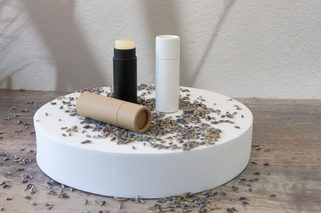 A soap and lip balm on a white round platform with lavender sprinkled around, on a wooden surface against a textured wall.
