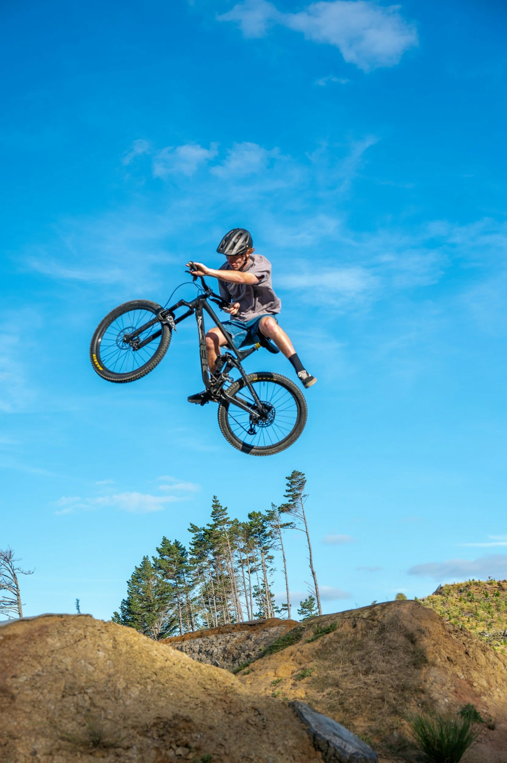 Mountain biker performing a jump on a dirt trail with clear blue sky and trees in the background.