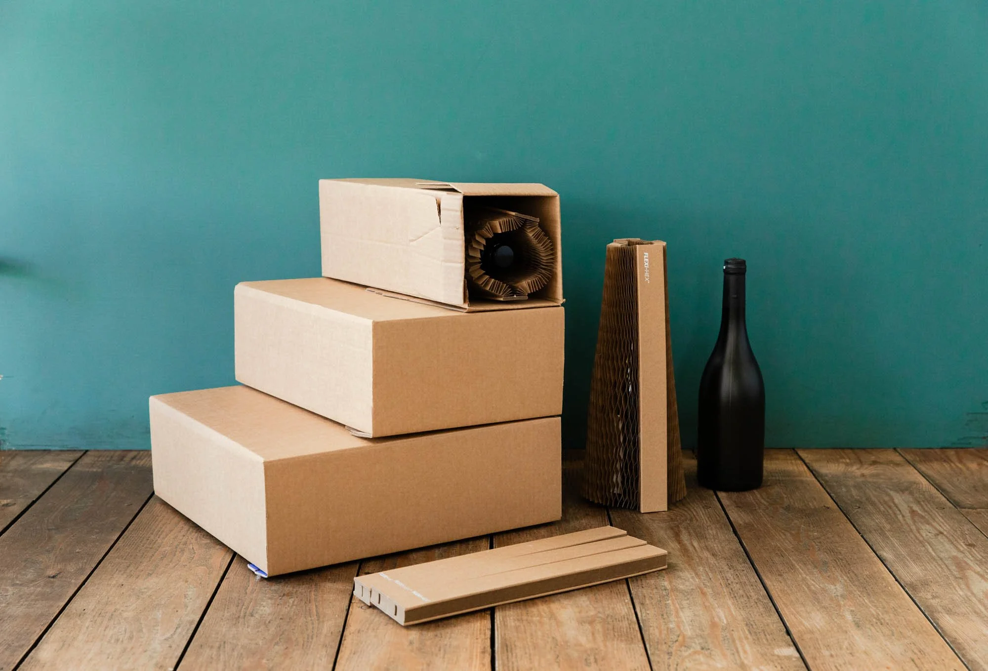 Cardboard boxes with packaging materials and a black wine bottle on a wooden floor.