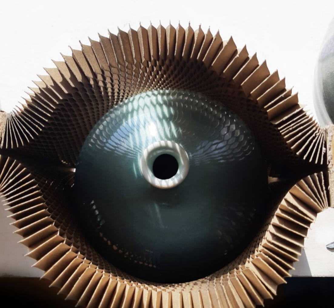 Large round ceramic vase with circular opening, partially encased in accordion-style cardboard packaging on white background.