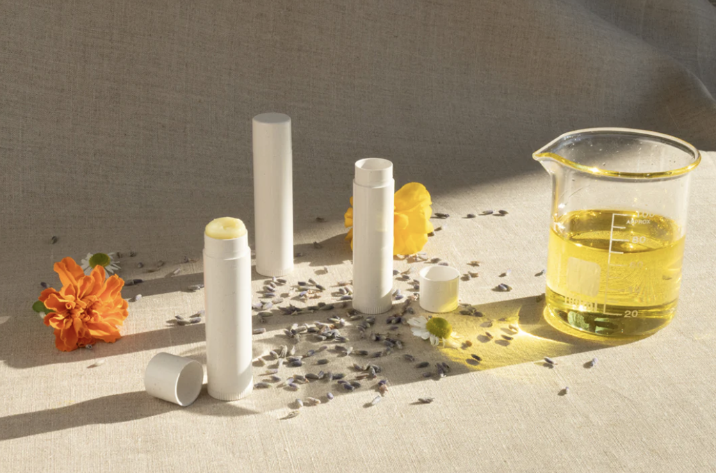 Lip balm tubes, a small container, a glass measuring cup with yellow liquid, and flowers on a light-colored surface