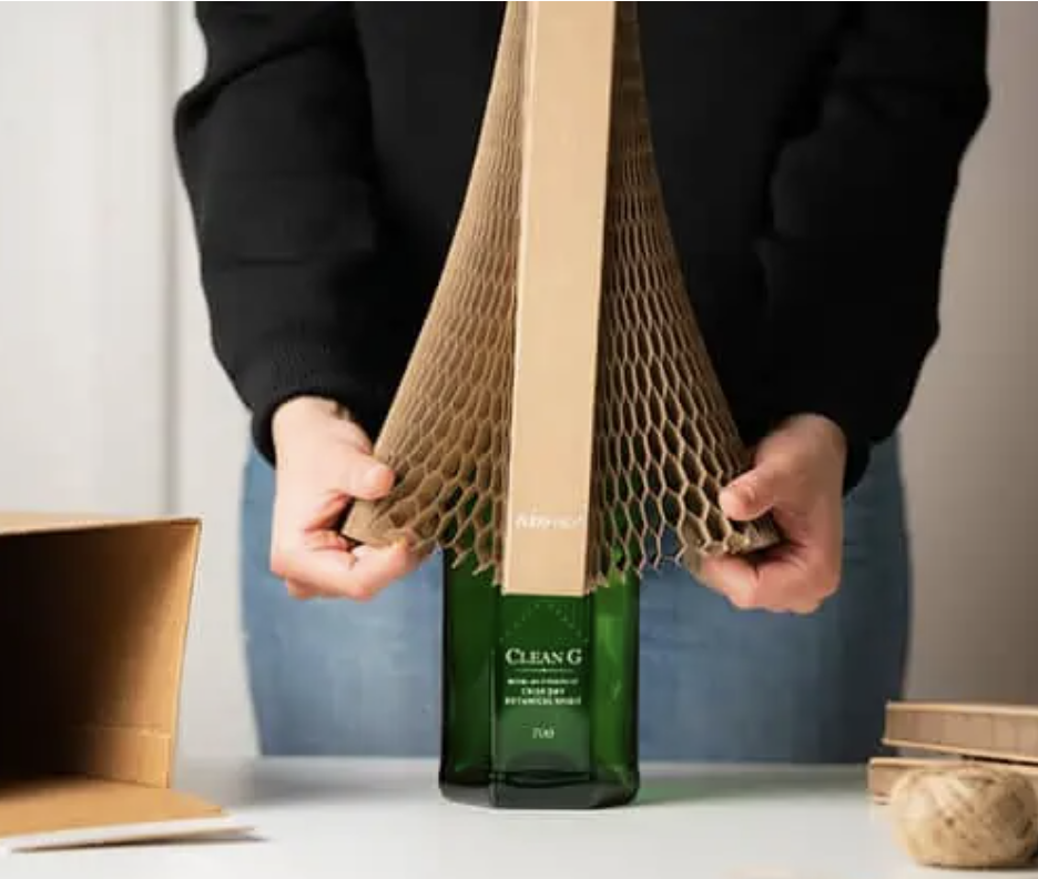 Person wrapping a green bottle in protective honeycomb cardboard packaging on a table.