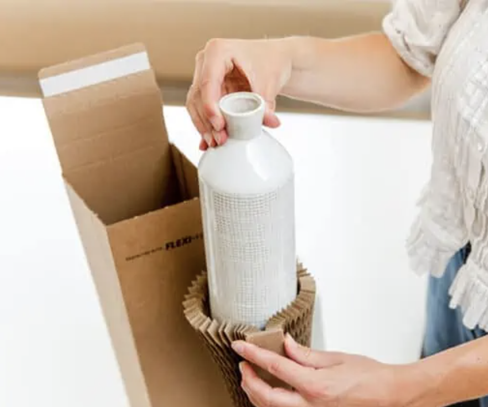 Person packaging a white ceramic vase with corrugated cardboard in a cardboard box.