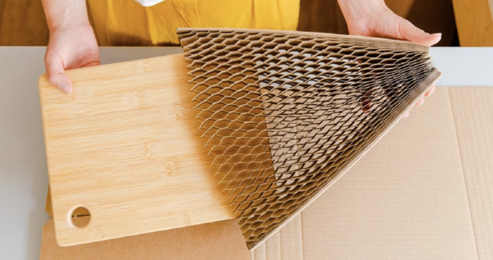 Person wrapping a bamboo cutting board in honeycomb cardboard packaging on a table.