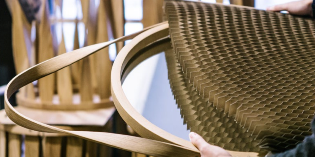 Person handling a flexible honeycomb cardboard sheet with wooden chair frames in the background.