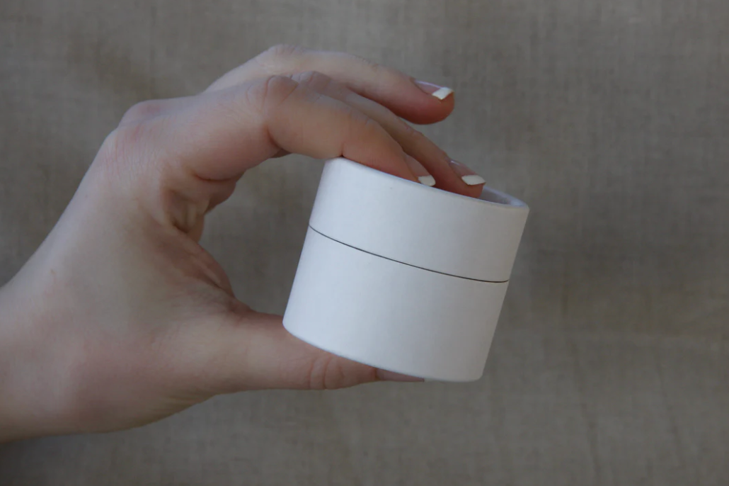 A hand with manicured nails holding a small, white, round container with a lid against a beige background.
