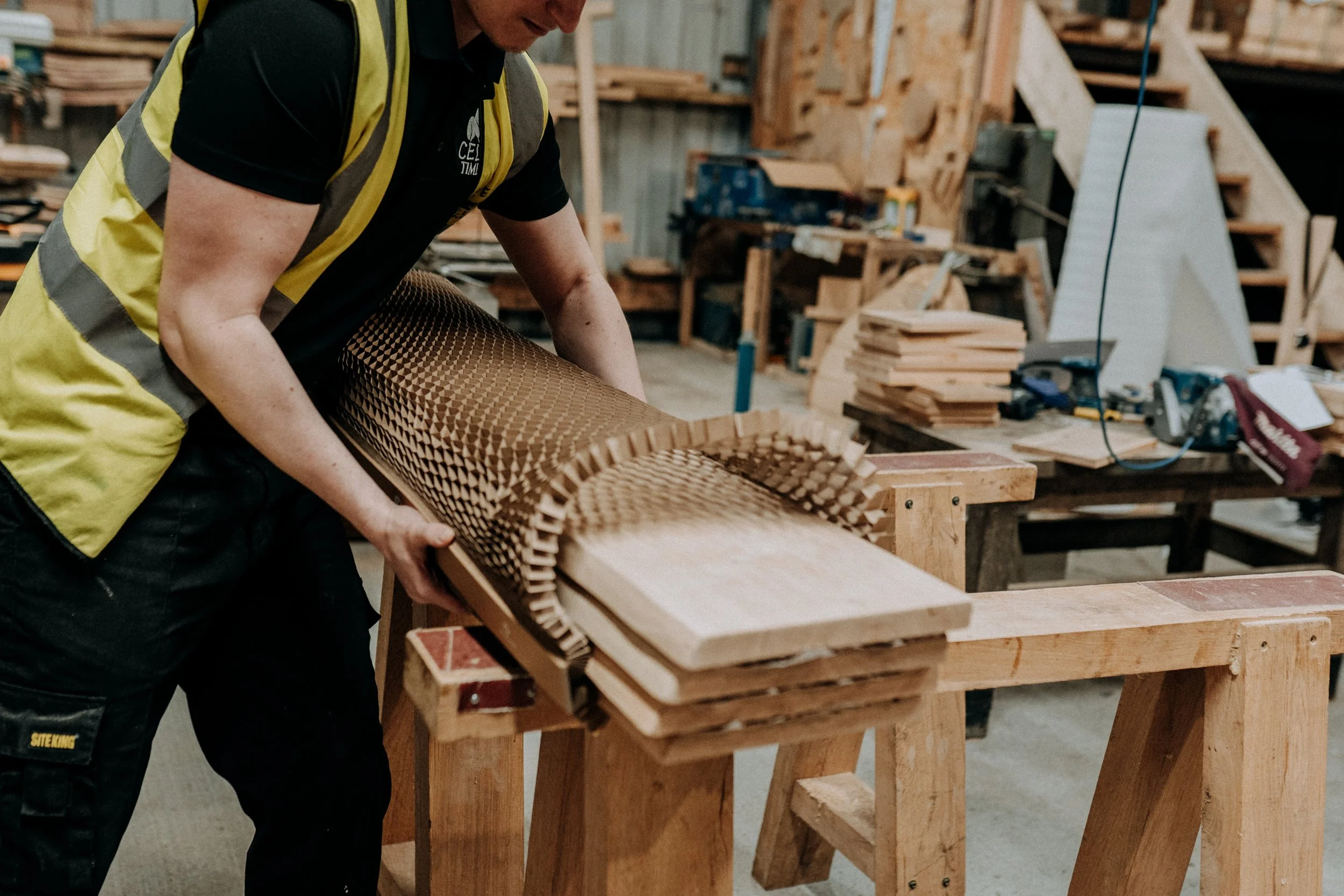 Person in high-visibility vest carrying wooden boards with a flexible accordion-like design in a carpentry workshop.