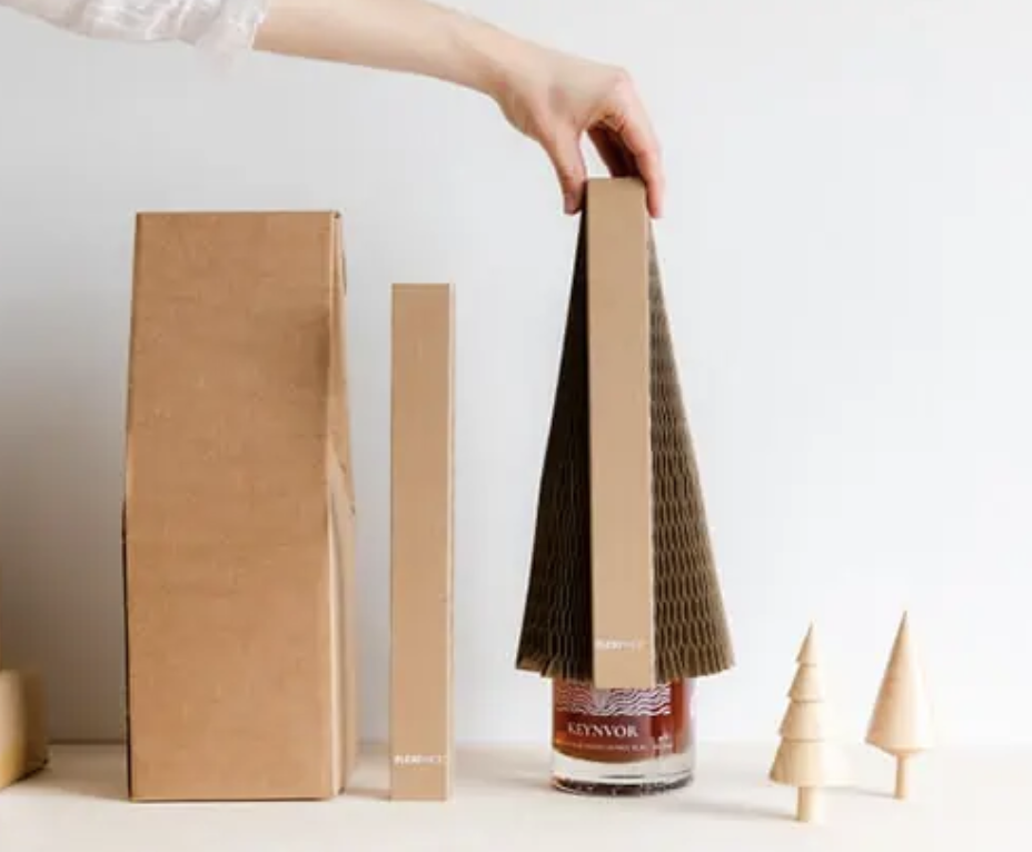 Brown cardboard packaging being placed over a glass bottle with a honeycomb sleeve, next to small wooden cone-shaped objects resembling trees, against a plain background.