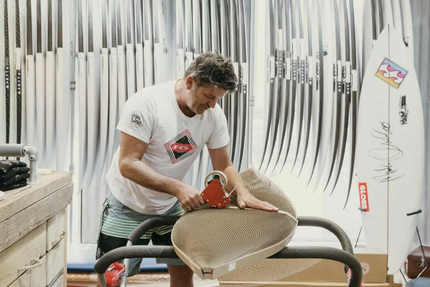 Man wrapping a surfboard in protective material, surrounded by surfboards.