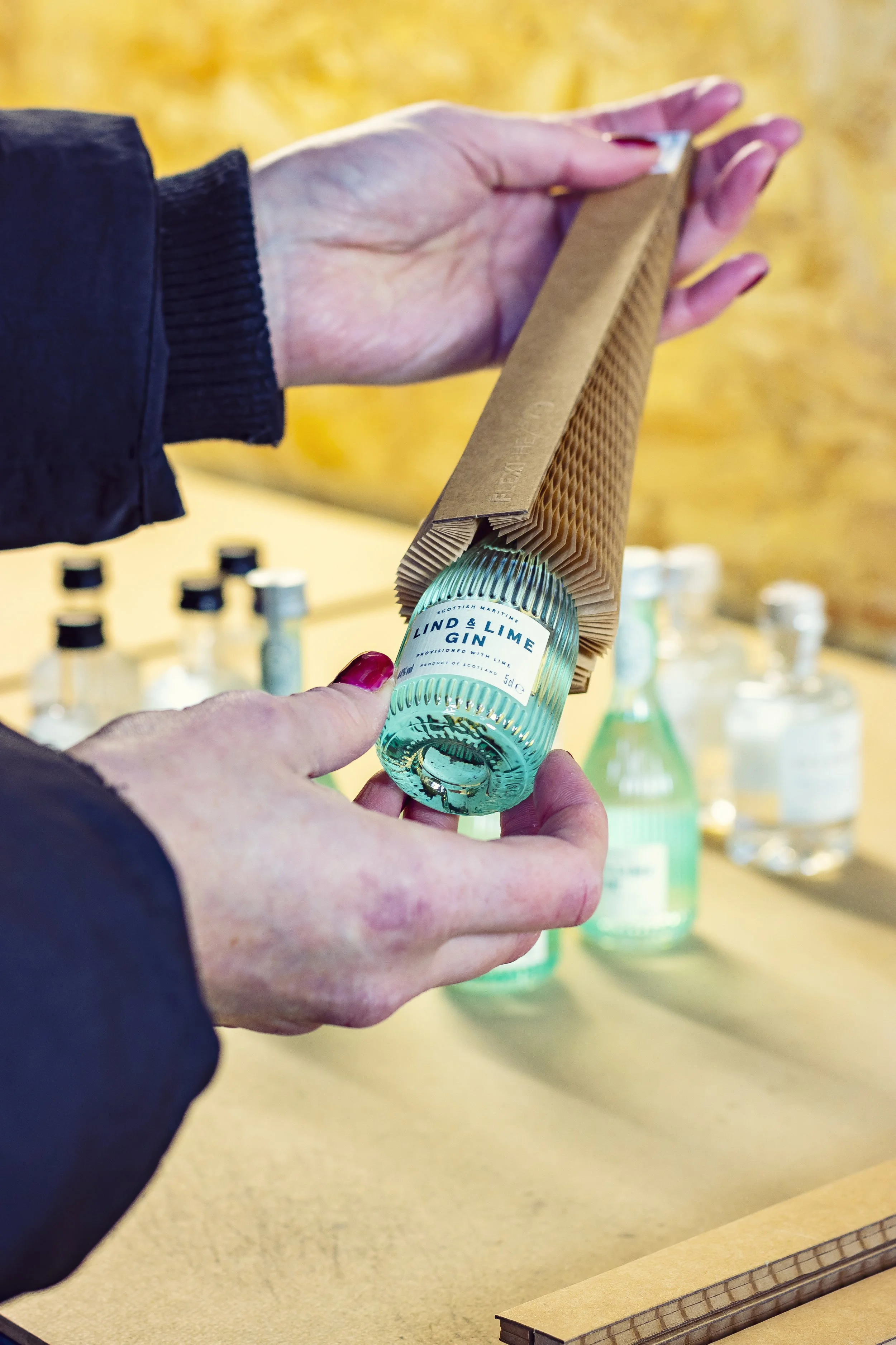 Person packaging a bottle of gin in a cardboard sleeve with more bottles in the background.