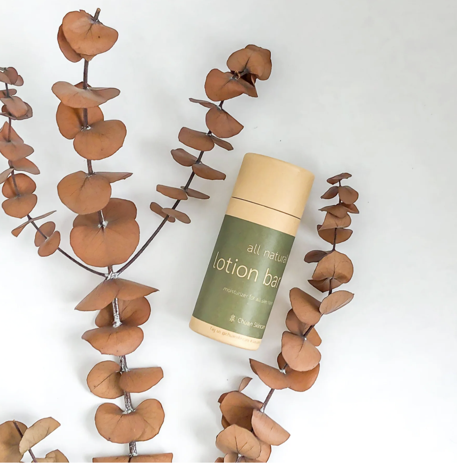 A green and beige container of all natural lotion bar surrounded by brown dried eucalyptus leaves on a white background.