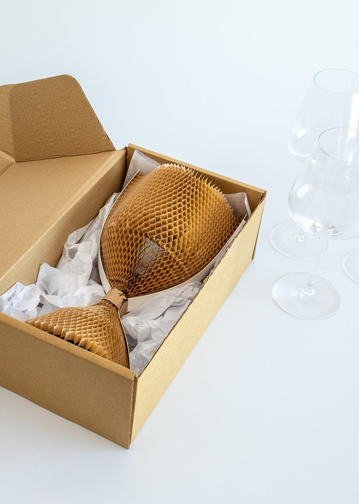 A cardboard box containing two wine glasses wrapped in honeycomb paper padding, with three empty glasses standing nearby.