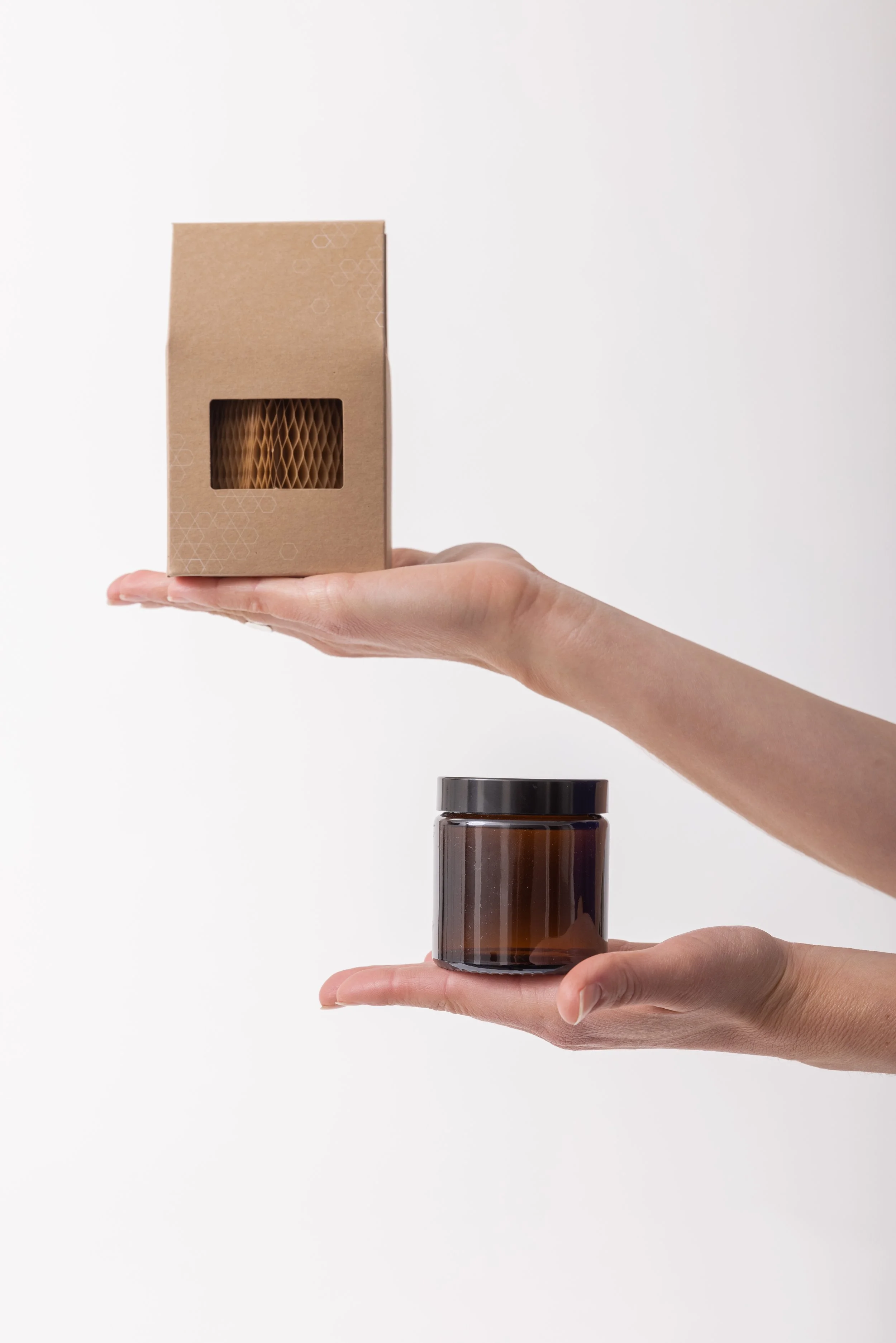 Two hands holding a brown cardboard box and a clear glass jar with a black lid, against a white background.