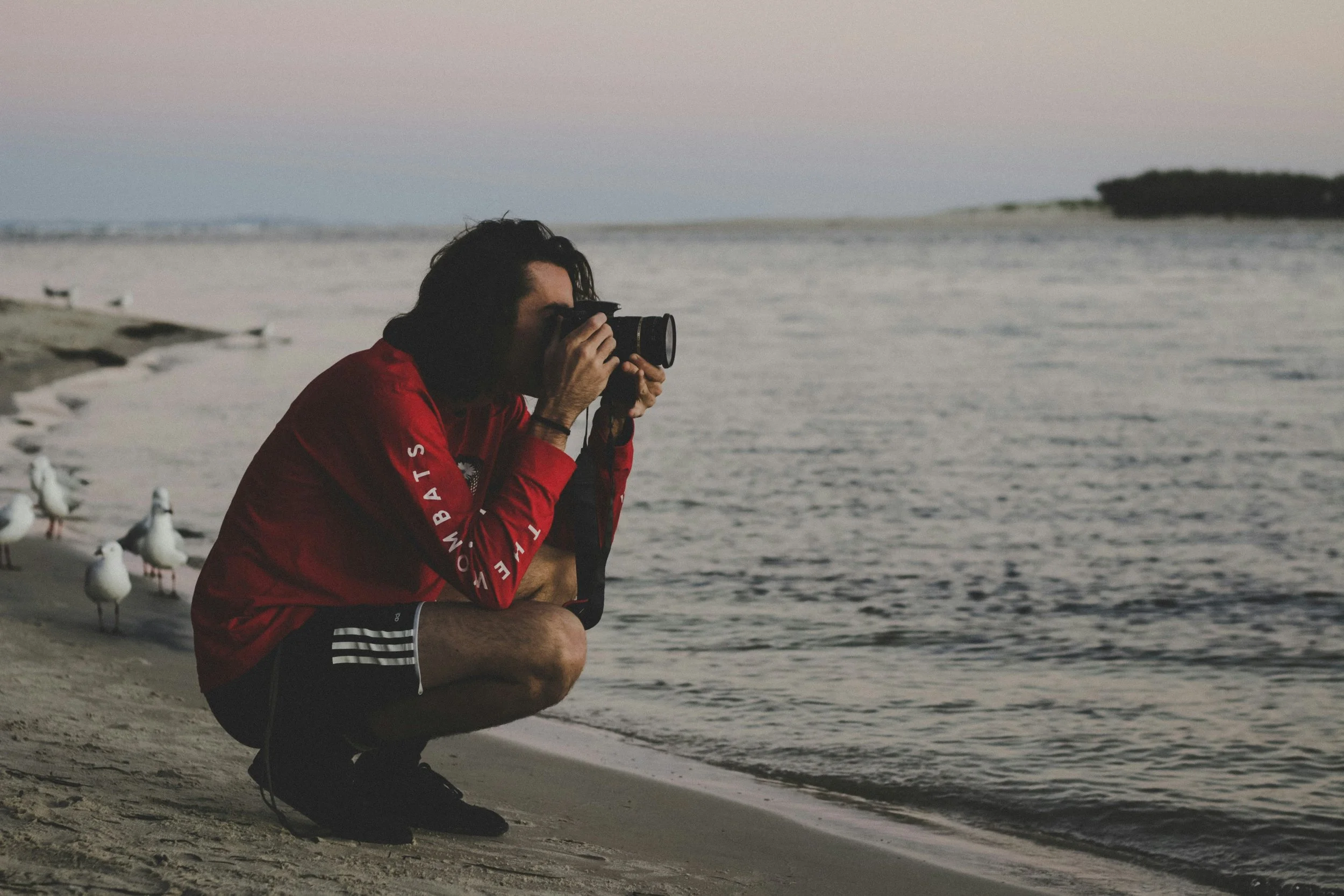 Photographer at the beach at dawn dressed in red Wombats top and shorts crouches at the shoreline to shoot image of the water.