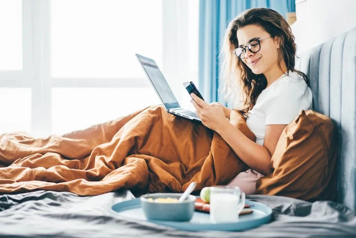 Happy woman with in bed working with phone and laptop