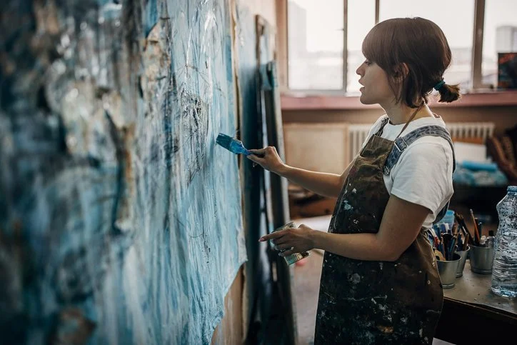 Woman adds paint to a large blue textured canvas painting.
