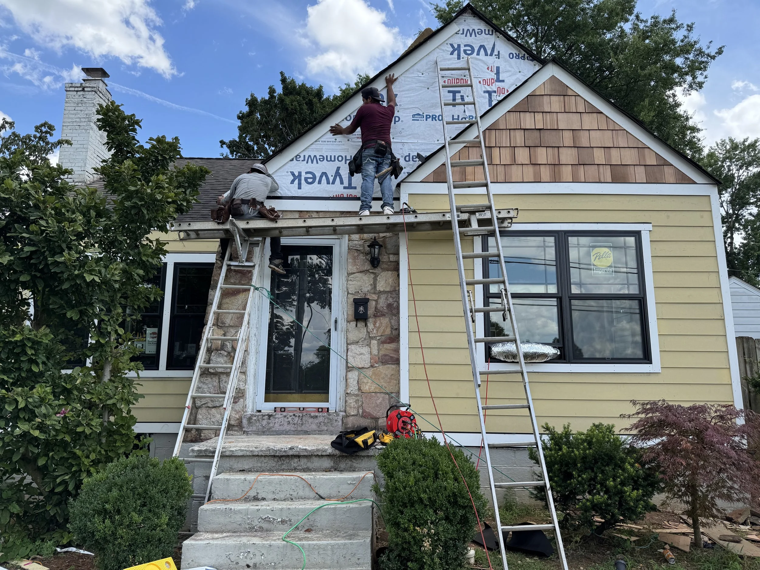 Two workers are on the roof of a house, installing new siding. One worker is standing and attaching siding, while the other is sitting on the roof. The house has yellow siding and large front windows, with ladders leaning against it. There are bushes and trees in the front yard, and construction tools are on the steps and ground.