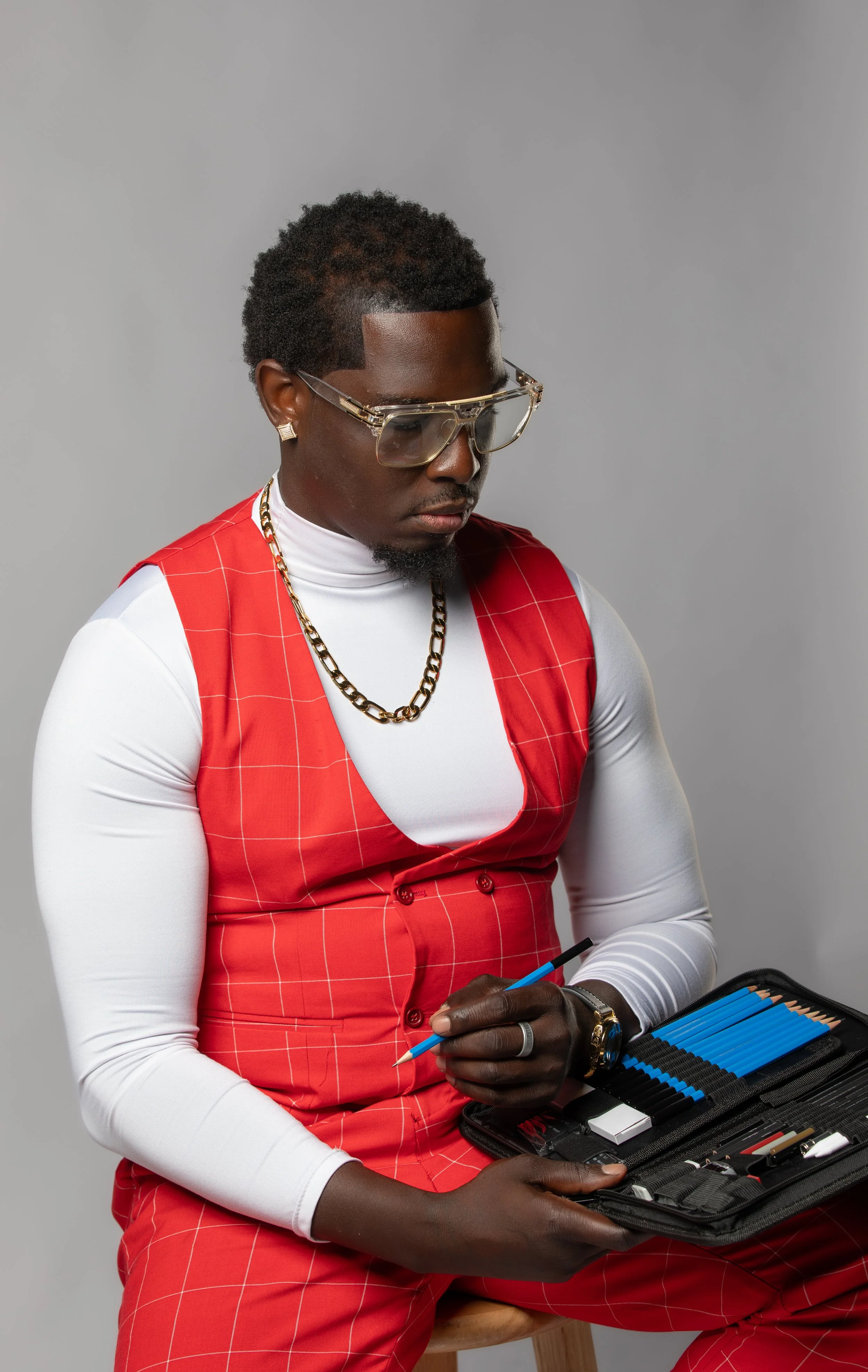 A man with dark skin and curly hair wearing glasses, a white turtleneck, a red checked vest, red checkered pants, gold jewelry, and white earrings, sitting on a wooden stool, holding a blue mechanical pencil, with an art case filled with colored pencils and markers on his lap against a plain gray background.