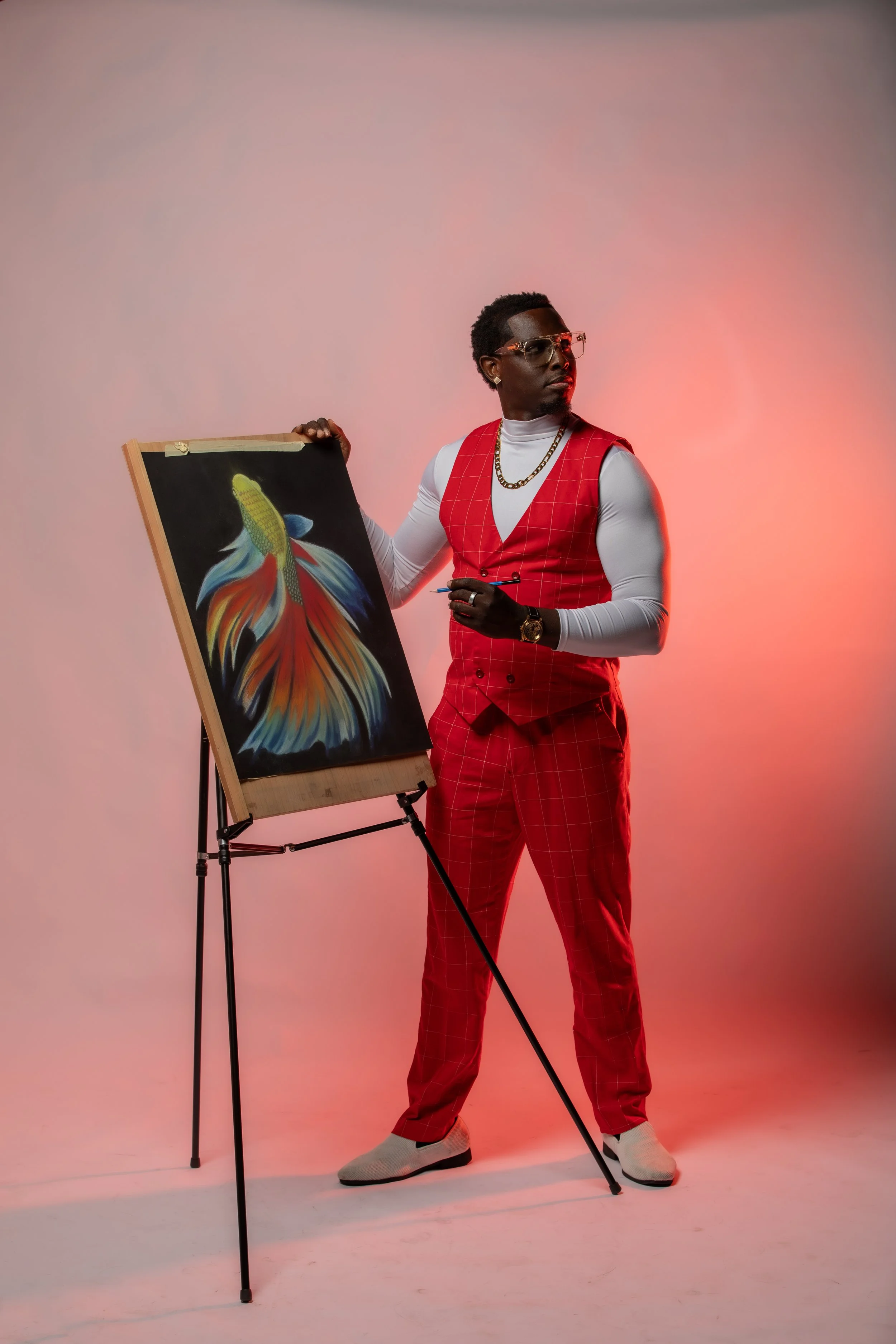 A man in a red checked suit, white turtleneck, and sunglasses stands next to an easel holding a painting of a colorful fish, with pink background lighting.