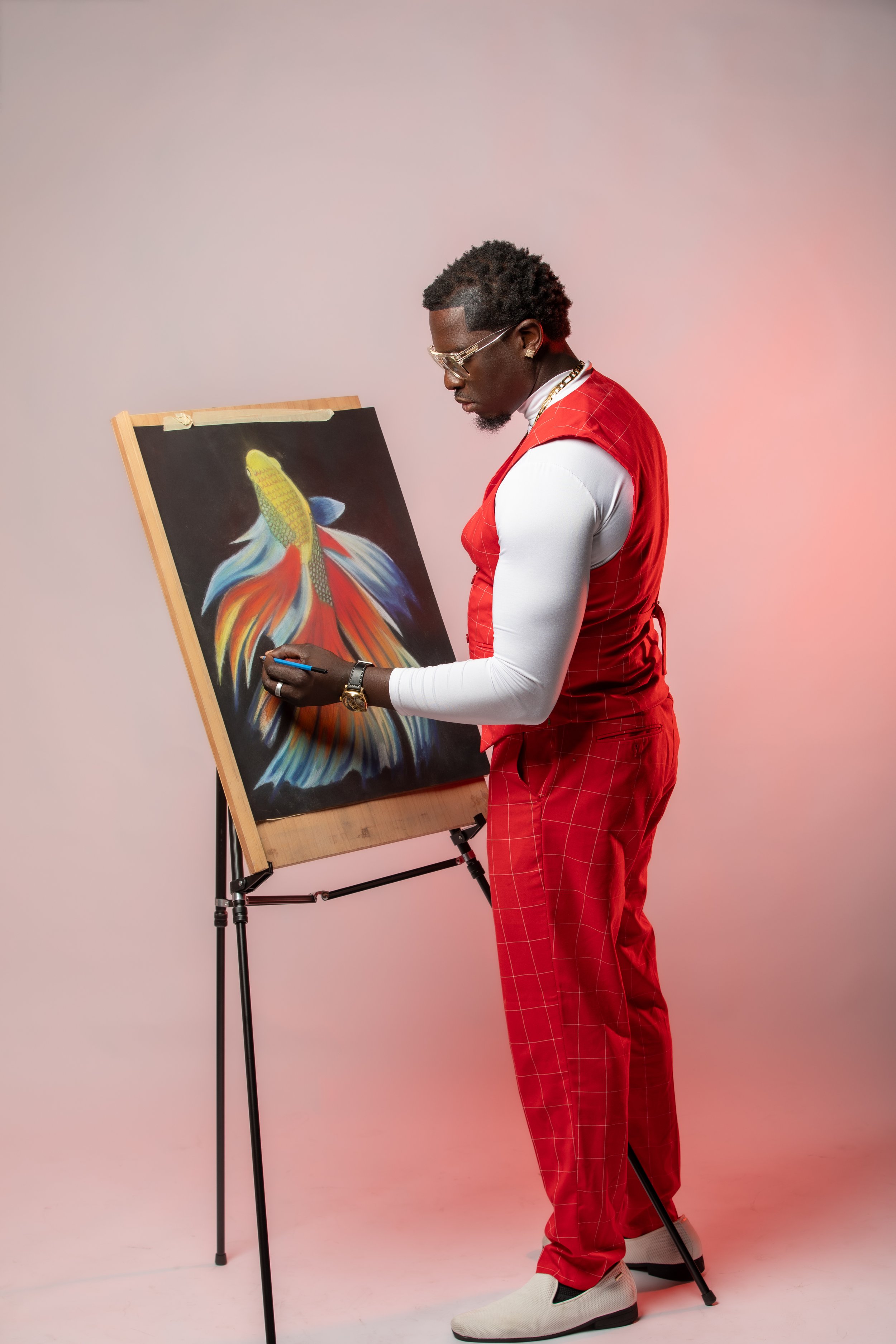 A man wearing red plaid pants, a white long sleeve shirt, with gold chains, glasses, and a watch, is standing and drawing or painting a colorful fish on a black canvas, which is mounted on an easel. The background is pinkish.