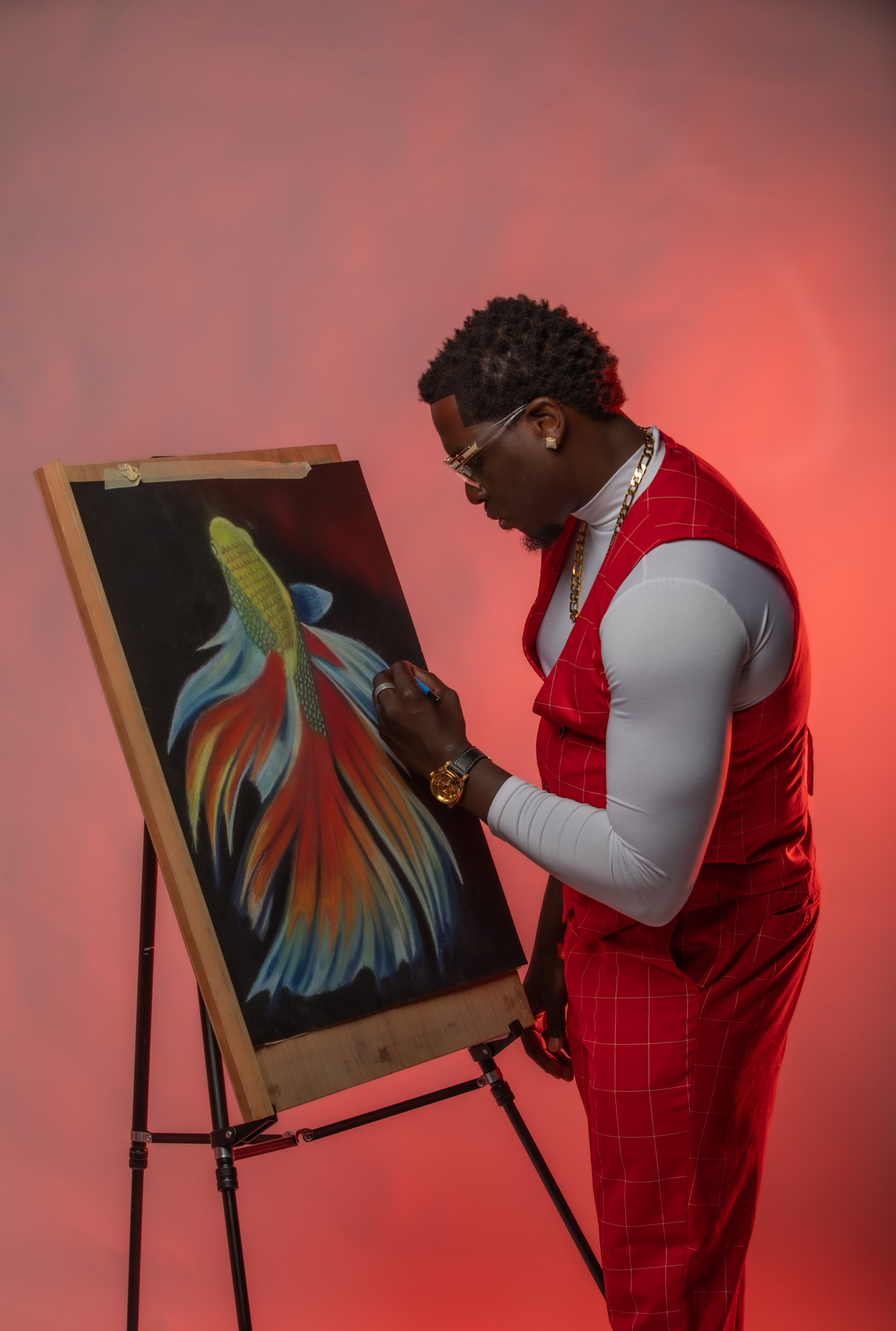 A man dressed in red plaid pants and a white shirt with a red vest, wearing glasses and gold jewelry, painting a vibrant, colorful fish on a canvas.