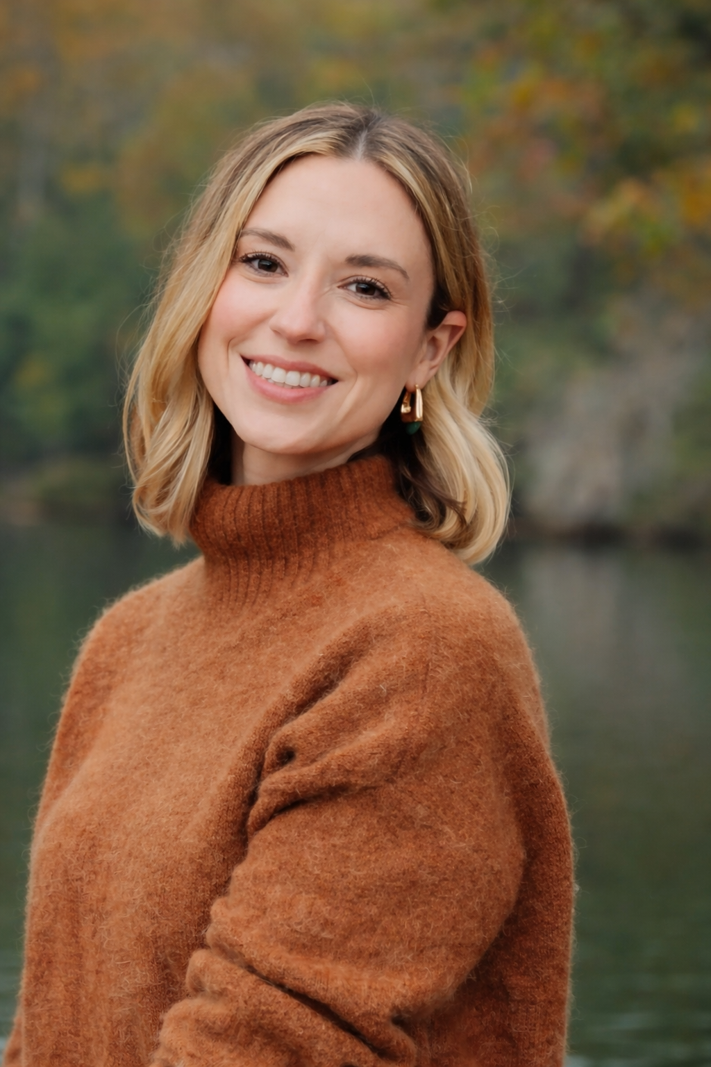 A smiling woman with shoulder-length blonde hair, wearing a brown turtleneck sweater, standing outdoors near a body of water with autumn trees in the background.