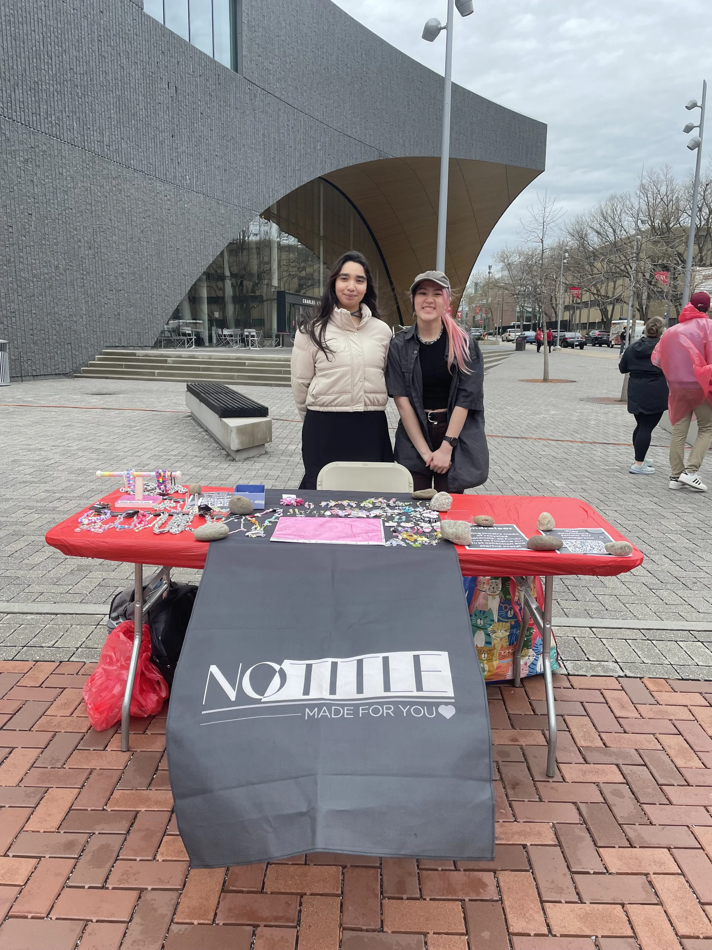 Two women standing behind a table with jewelry and accessories outside of a modern building, with people walking in the background while it rains. On the left is cofounder Isel Jimenez and on the right is founder Samantha Salomon.