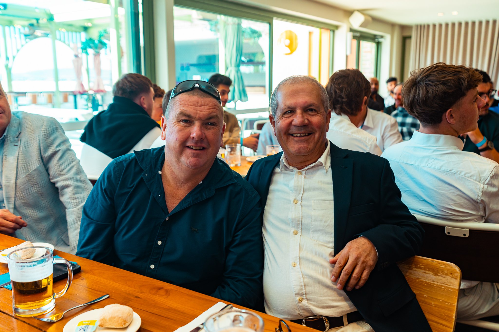 Two men sitting side by side at a table in a lively restaurant or pub, smiling and posing for the photo. The man on the left is wearing a dark shirt with sunglasses on his head, and the man on the right is dressed in a white shirt with a dark blazer.