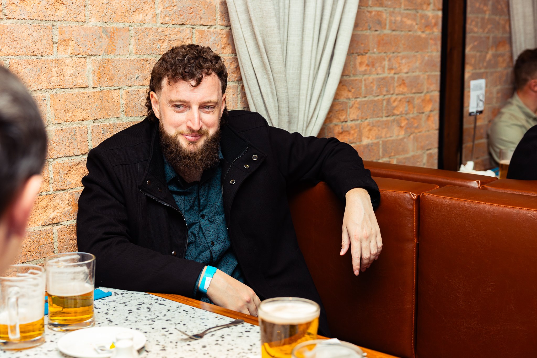 A man with curly hair and a beard sitting at a restaurant table with beer glasses, looking at someone off-camera, against a brick wall background.