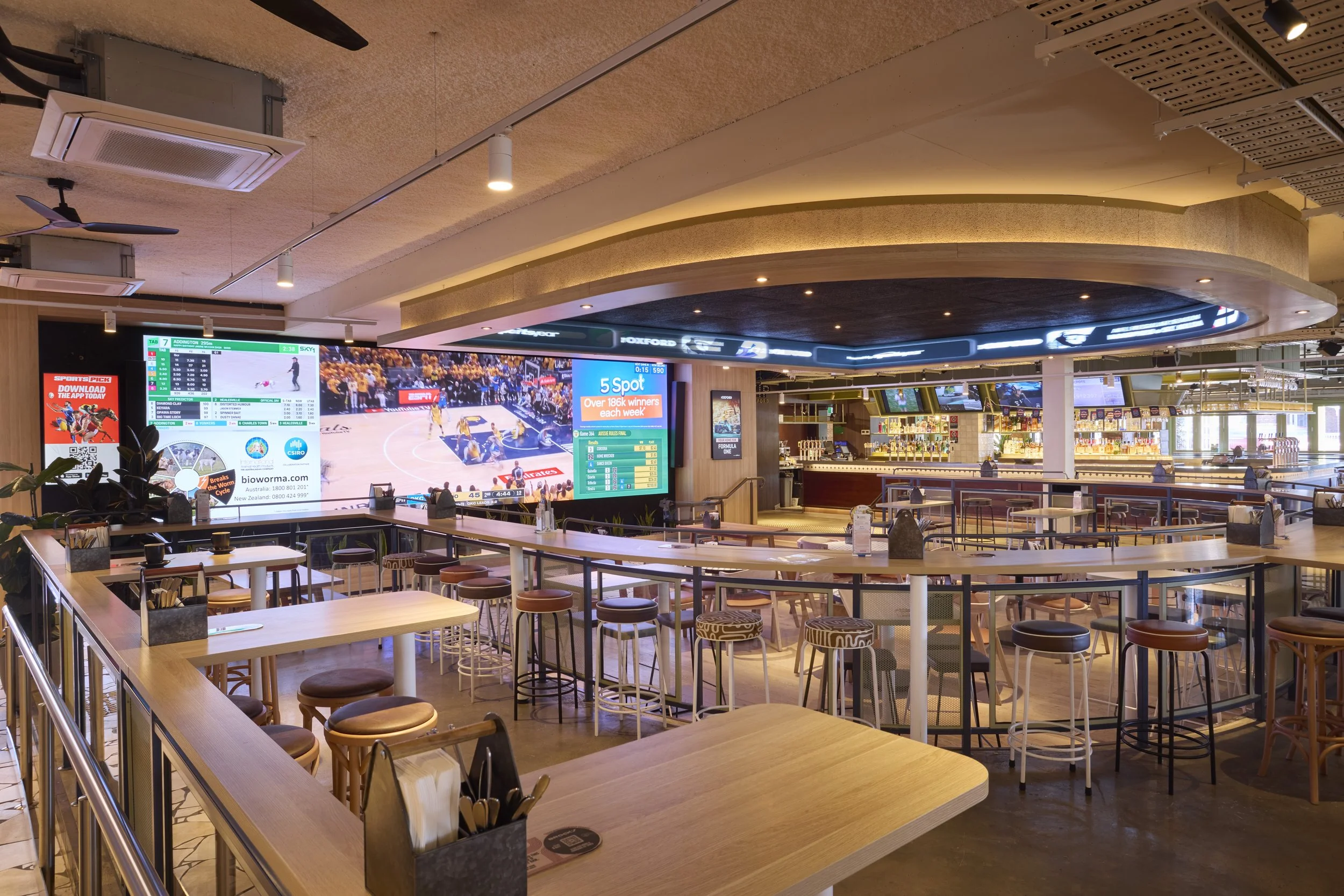 Interior view of a sports bar or lounge with multiple television screens showing a basketball game, seating areas with tables and chairs, and a bar counter in the background.