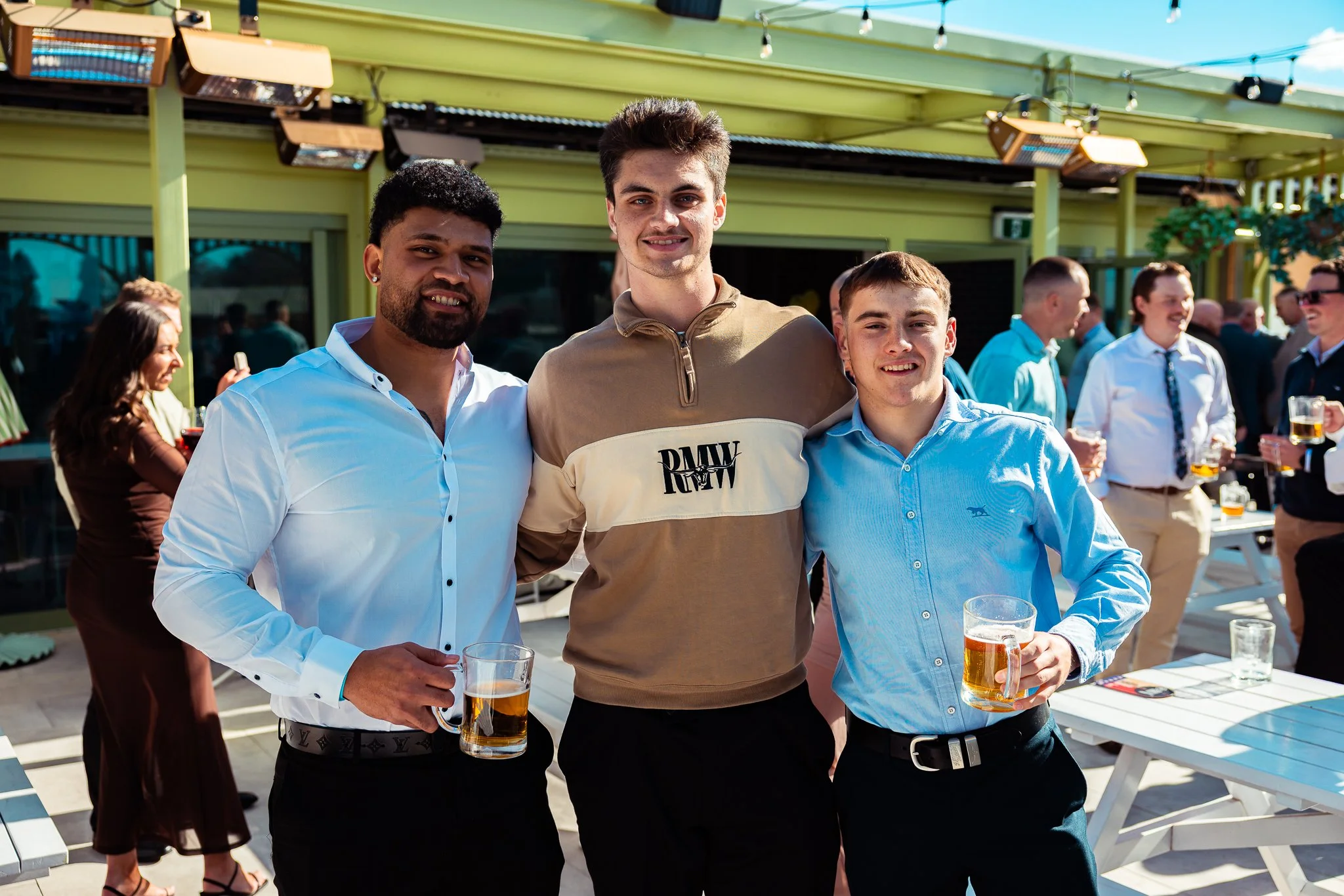 Three young men at an outdoor gathering, holding glasses of beer, smiling at the camera.