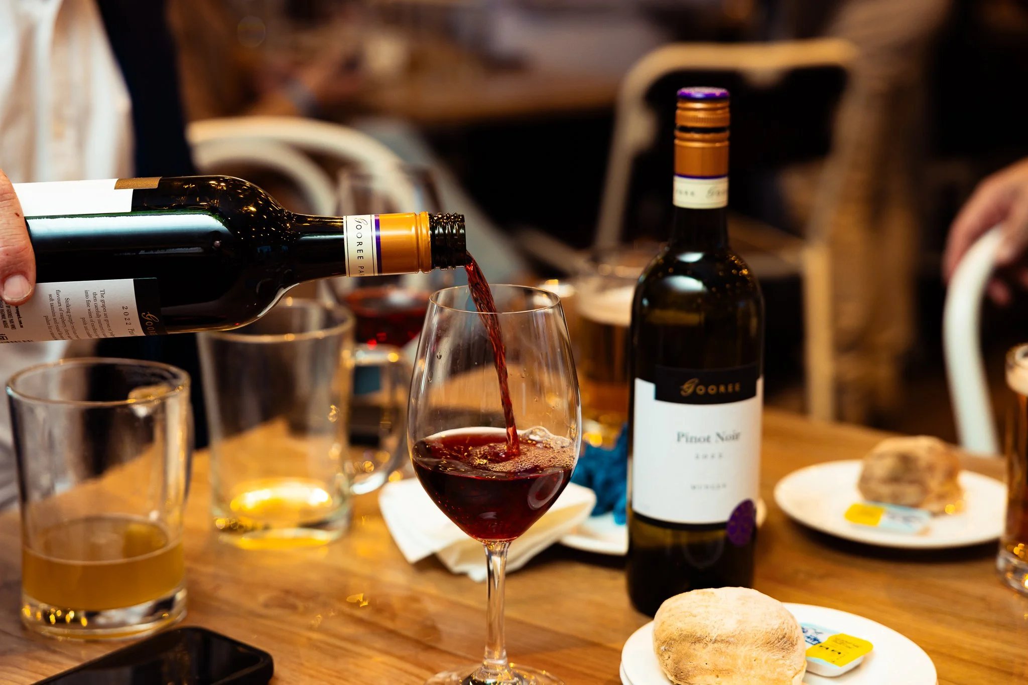 A person pouring red wine into a glass at a gathering or dinner party with other drinks and food on the table.