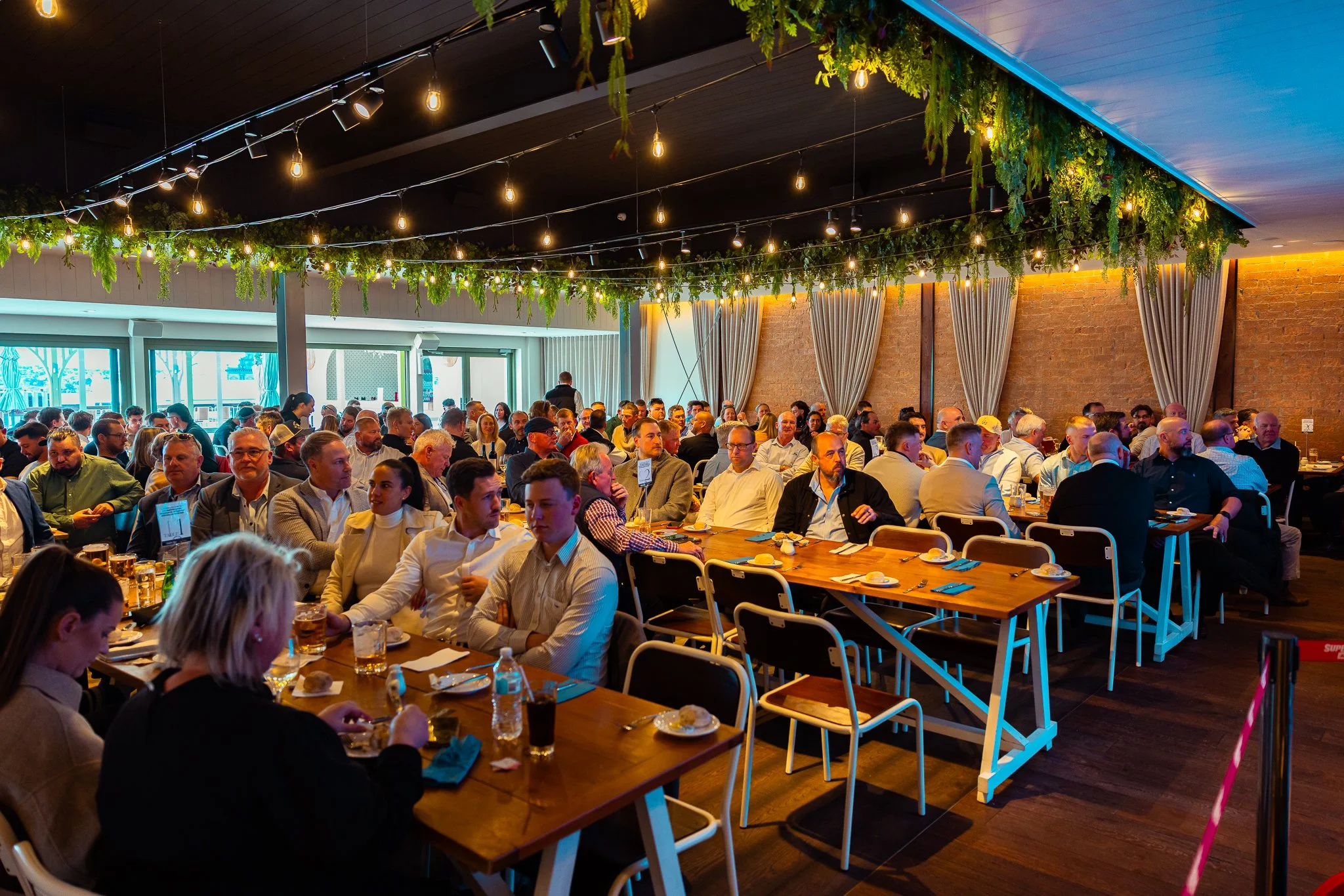 People gathered at a large indoor event space with high ceilings decorated with hanging string lights and greenery. The attendees are seated at long wooden tables with place settings, including plates, glasses, and cutlery. There are large windows an