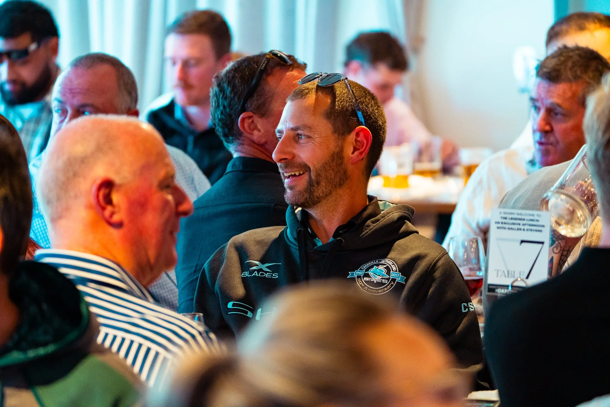 A group of men sitting at a table during a social event. The man in the foreground is smiling and wearing glasses on his head, with a dark polo shirt that has a sports team logo. Others are engaged in conversation, with some partially visible. The se