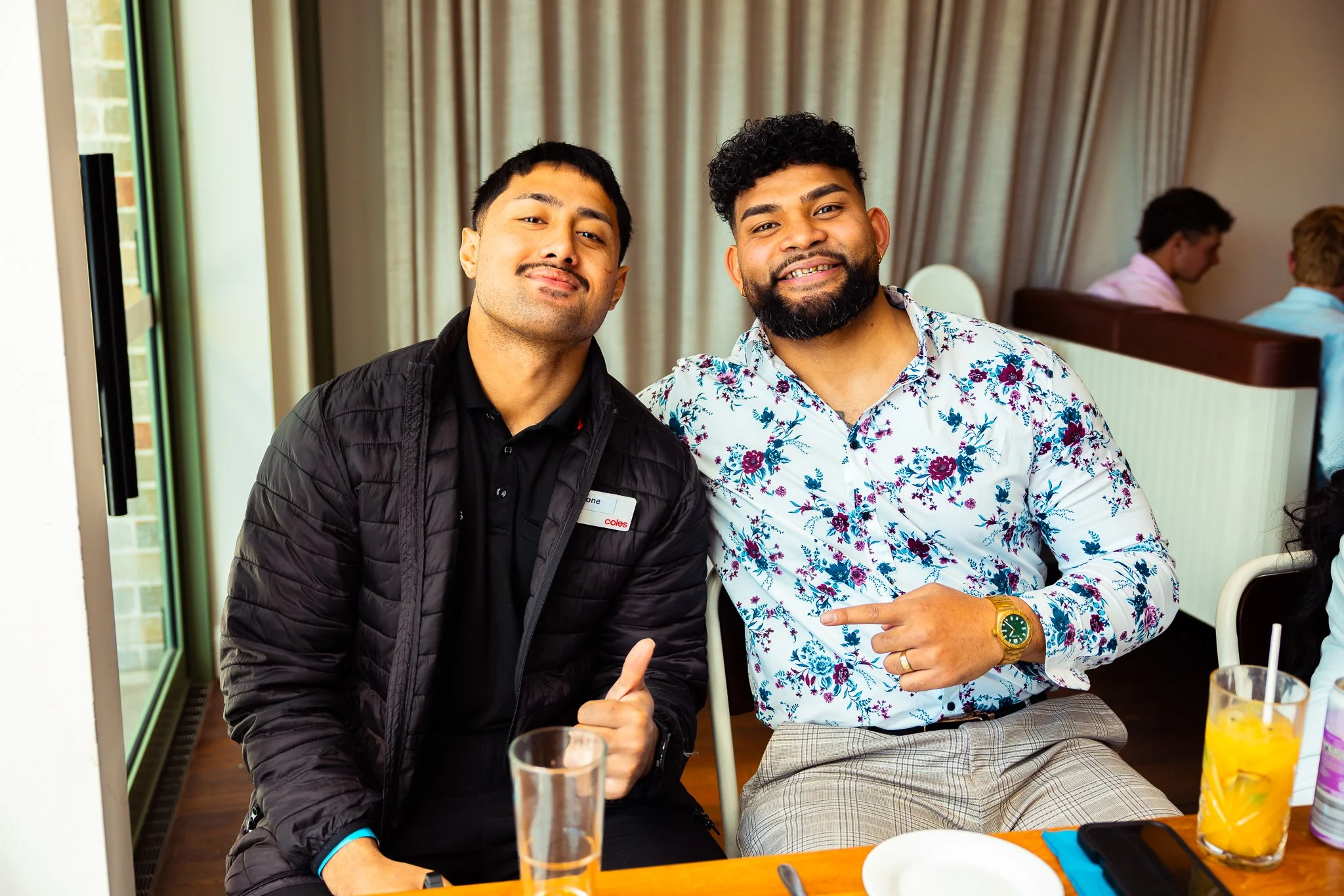 Two men sitting together at a table, smiling at the camera, with drinks and snacks on the table, in a room with a window and curtains in the background.