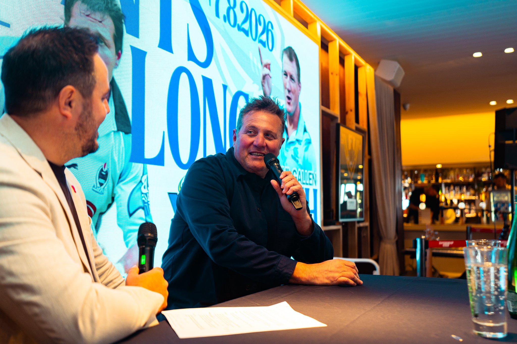 Two men participating in a panel or discussion at an event, with a large screen displaying a graphic in the background. One man is speaking into a microphone, while the other is looking down at notes.