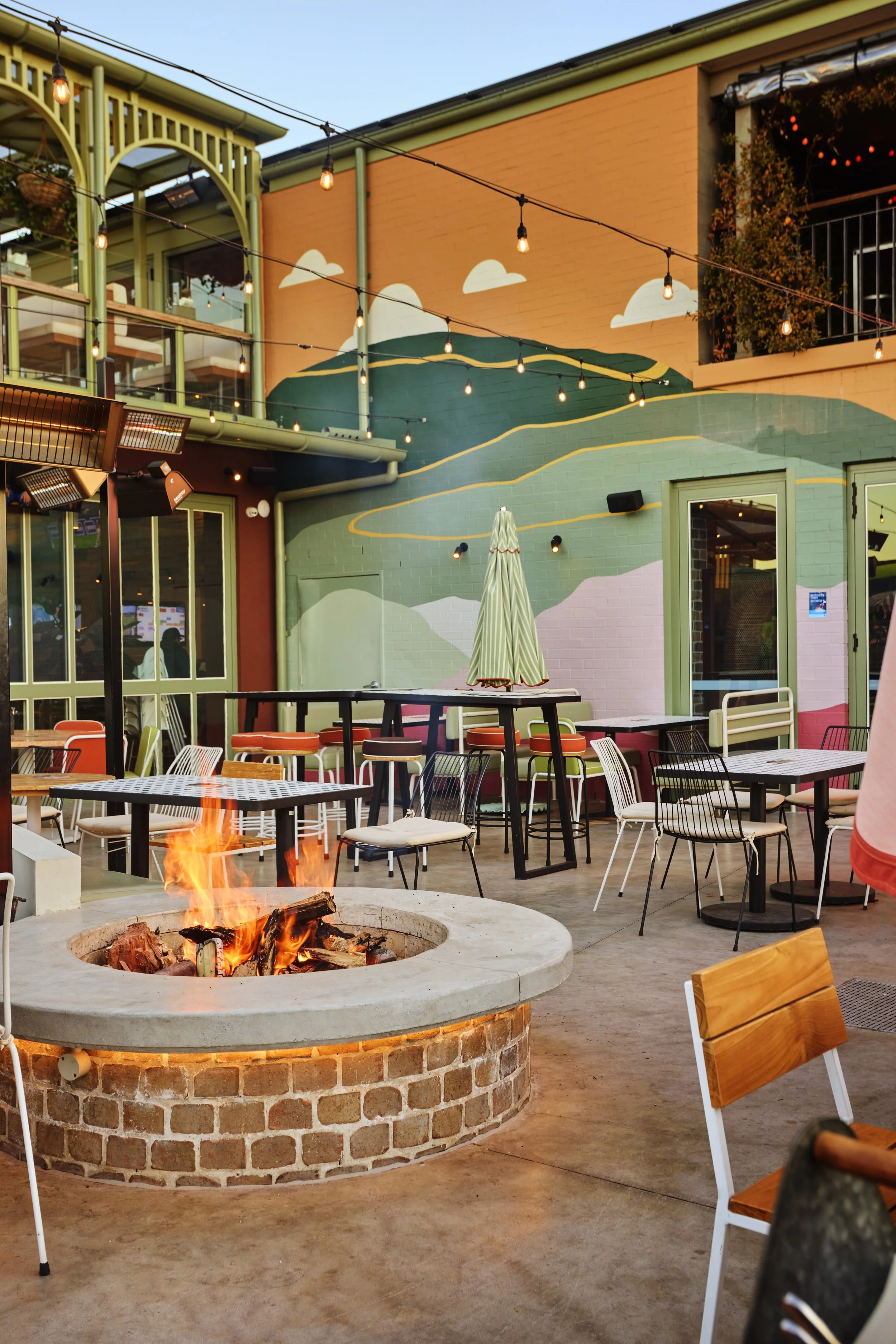 Outdoor patio area with tables, chairs, a fire pit, string lights, and colorful mural on the wall.