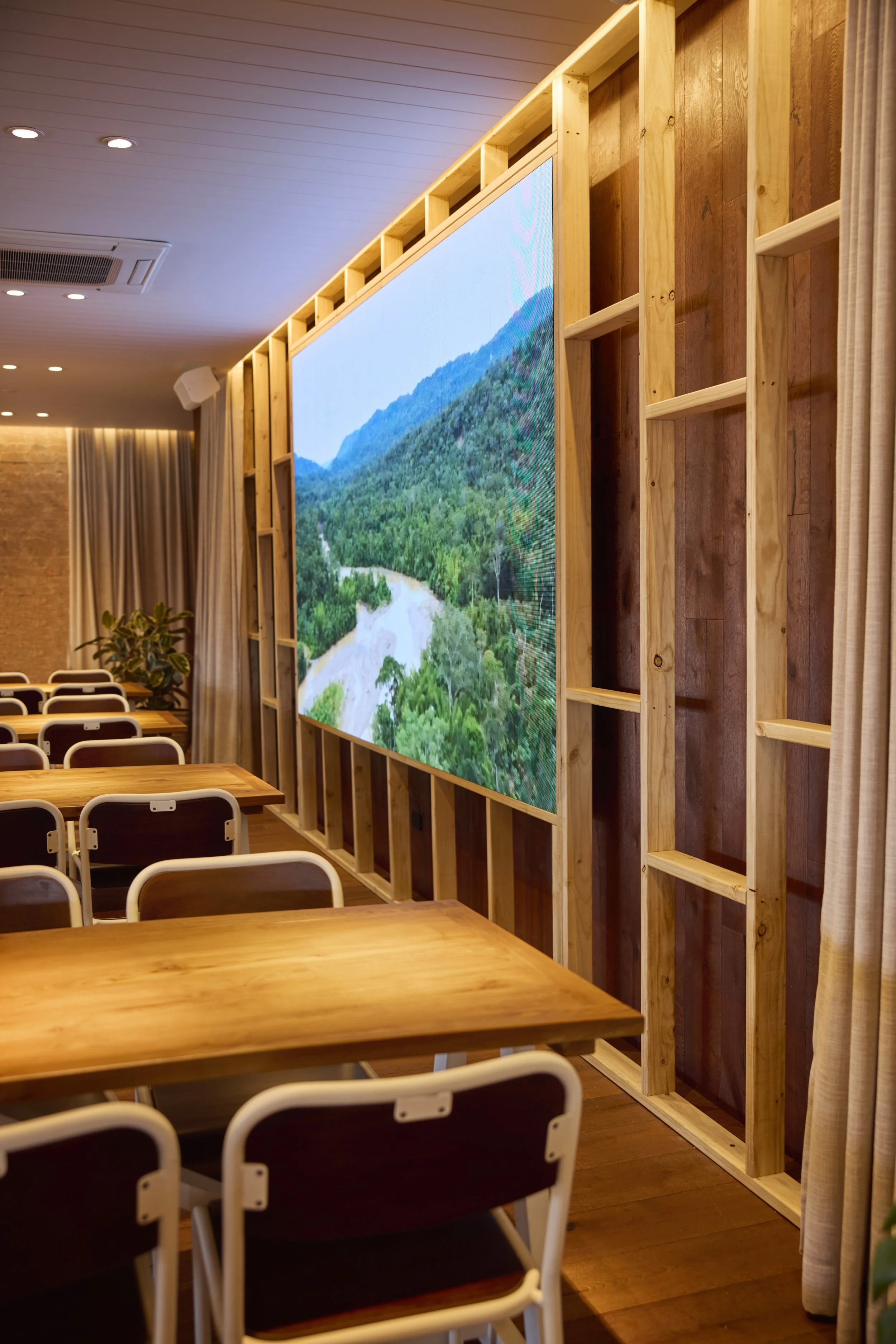 Indoor space with wooden tables and chairs, a large digital screen displaying a nature scene of mountains and forest, and a wooden shelving unit on the wall.