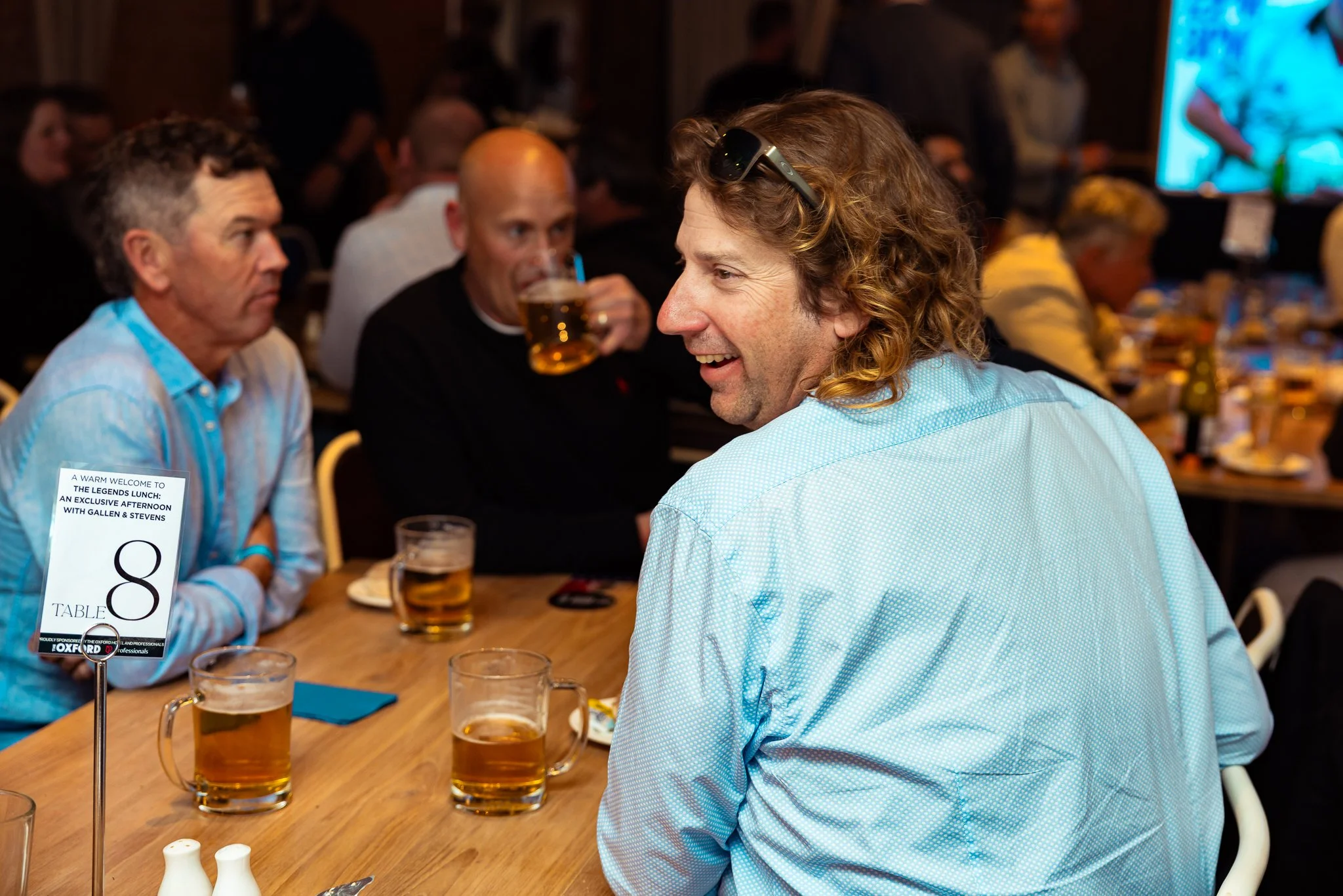 A man with curly hair and sunglasses on his head smiling and talking to others in a crowded restaurant. Several glasses of beer and a table number sign are visible.