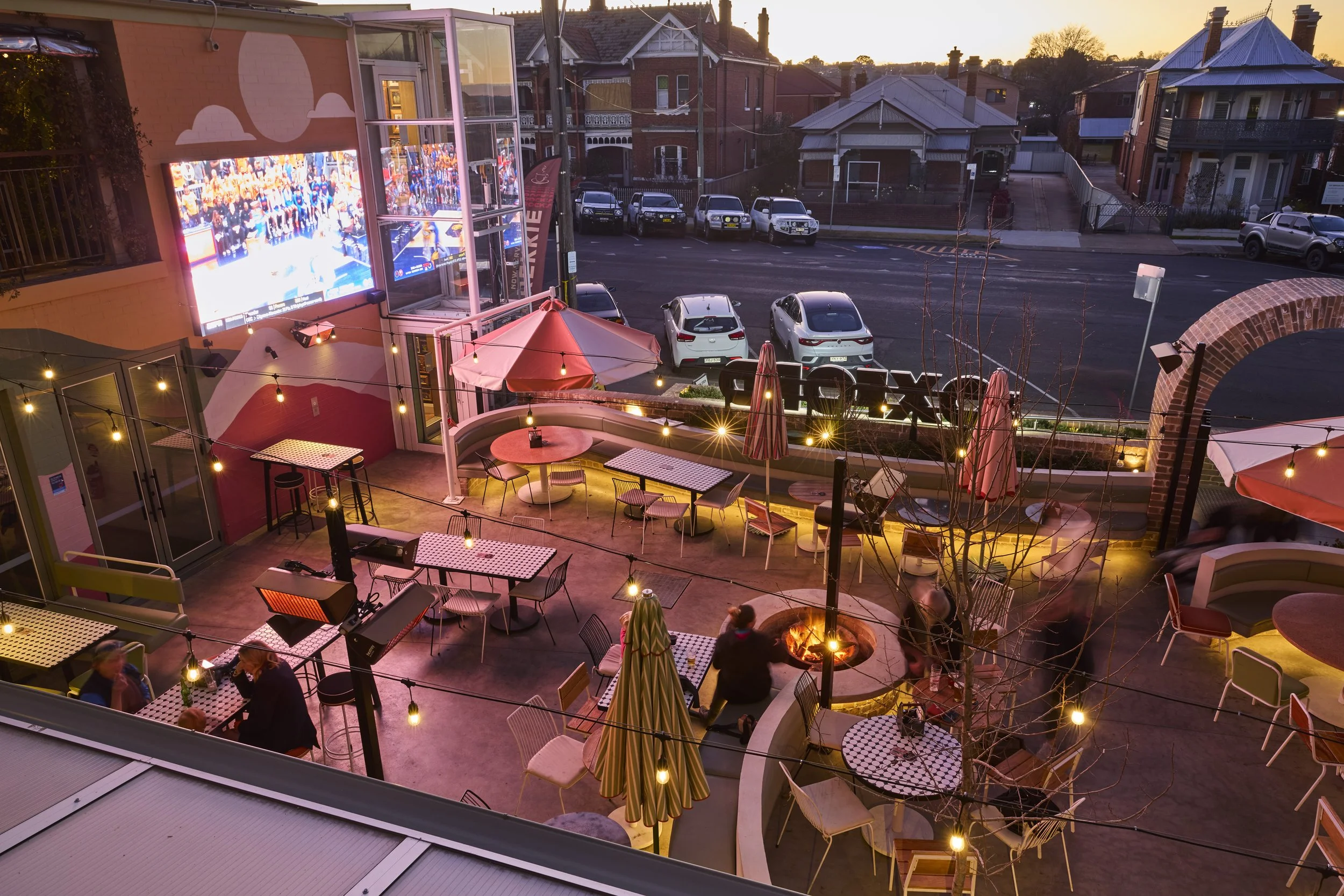 Outdoor patio of a restaurant or bar during evening with string lights, tables, and umbrellas. Several cars are parked across the street, with residential houses in the background, and a large outdoor TV screen displaying a sports event.