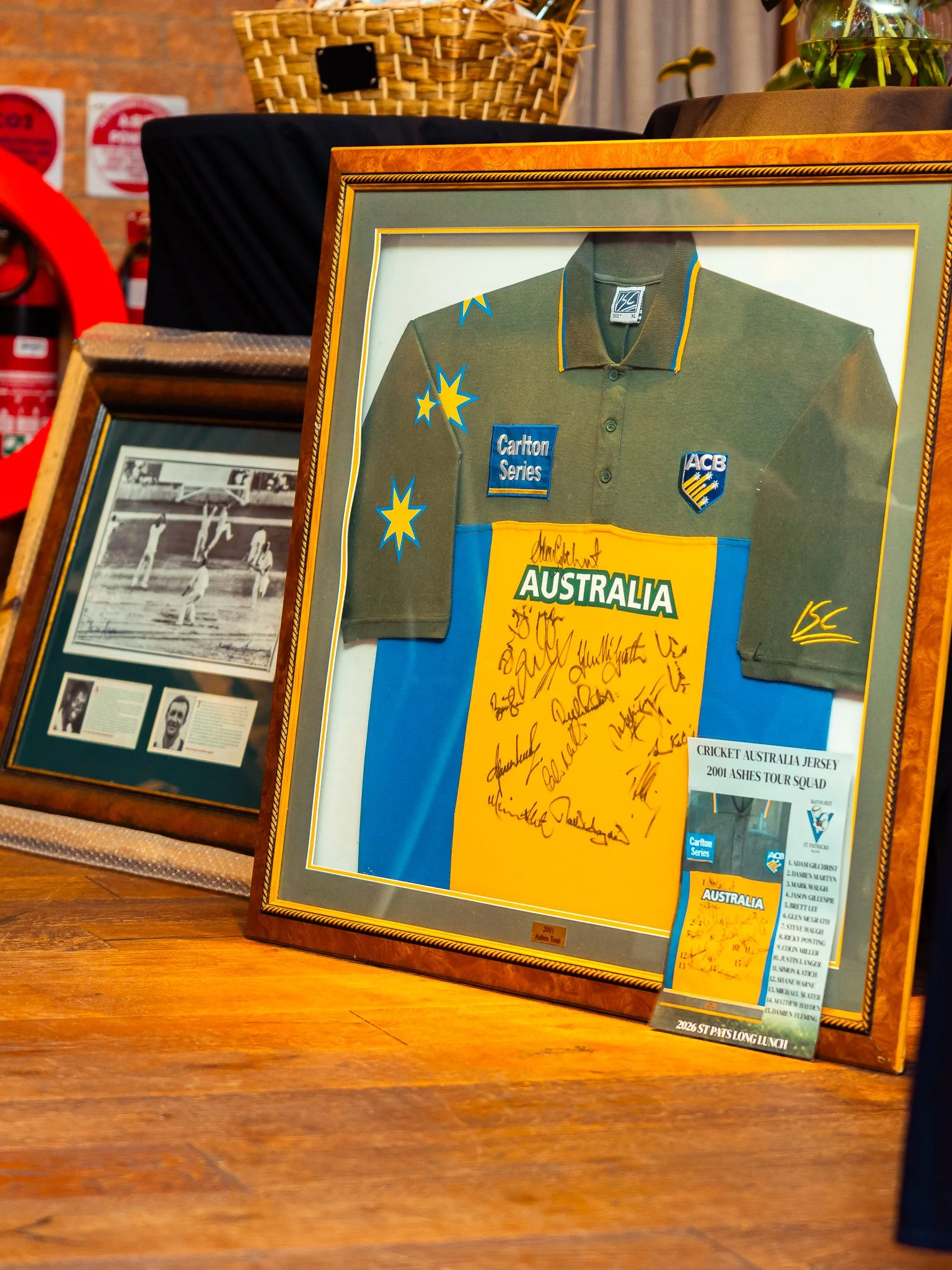 A framed cricket jersey from Australia with autographs, displayed on a wooden surface, with a black and white photograph and other objects in the background.