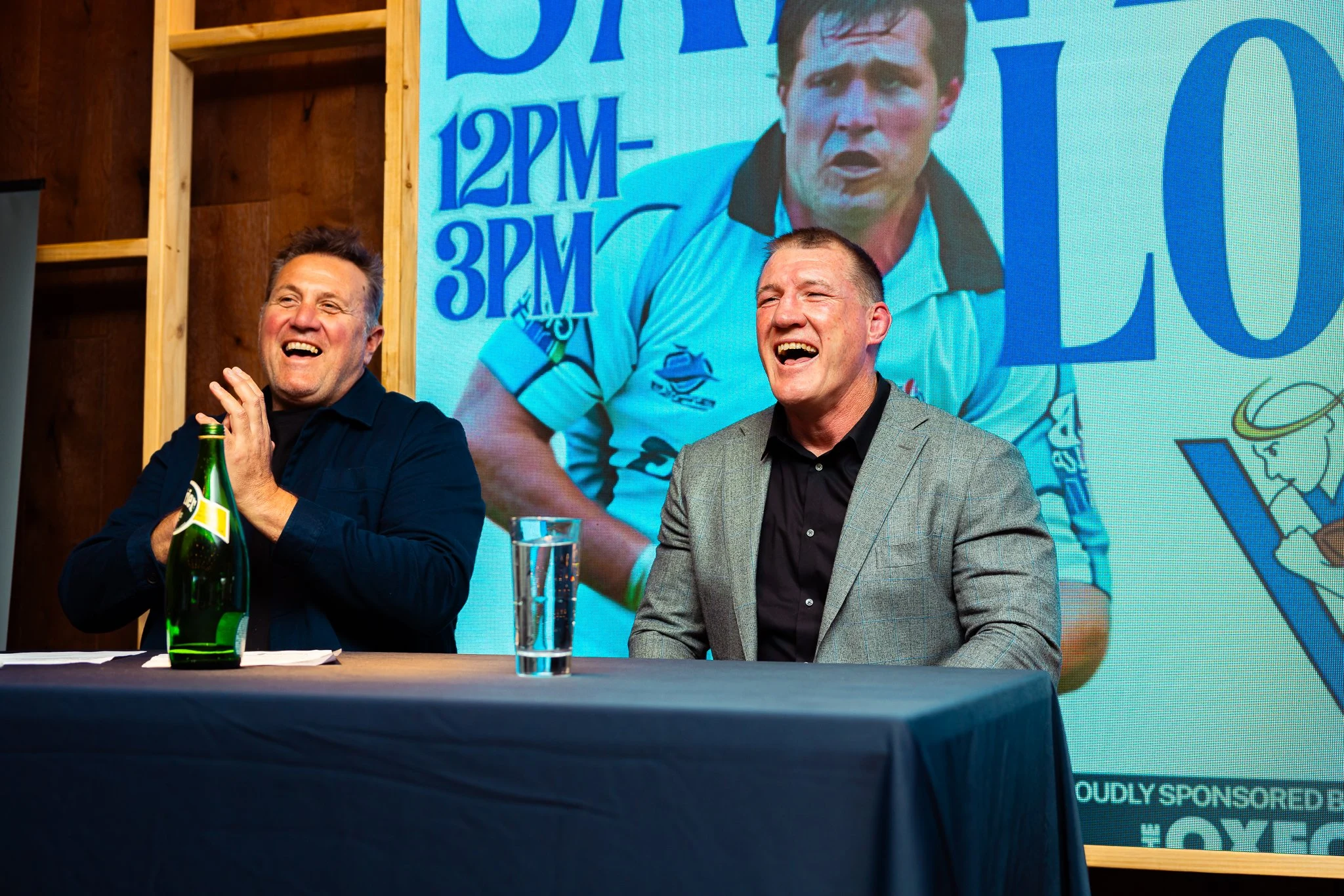 Two men sitting at a table during an event, one laughing and holding a green wine bottle, the other smiling with a glass of water in front of him. Behind them, a large screen displays a sports figure and advertising text.