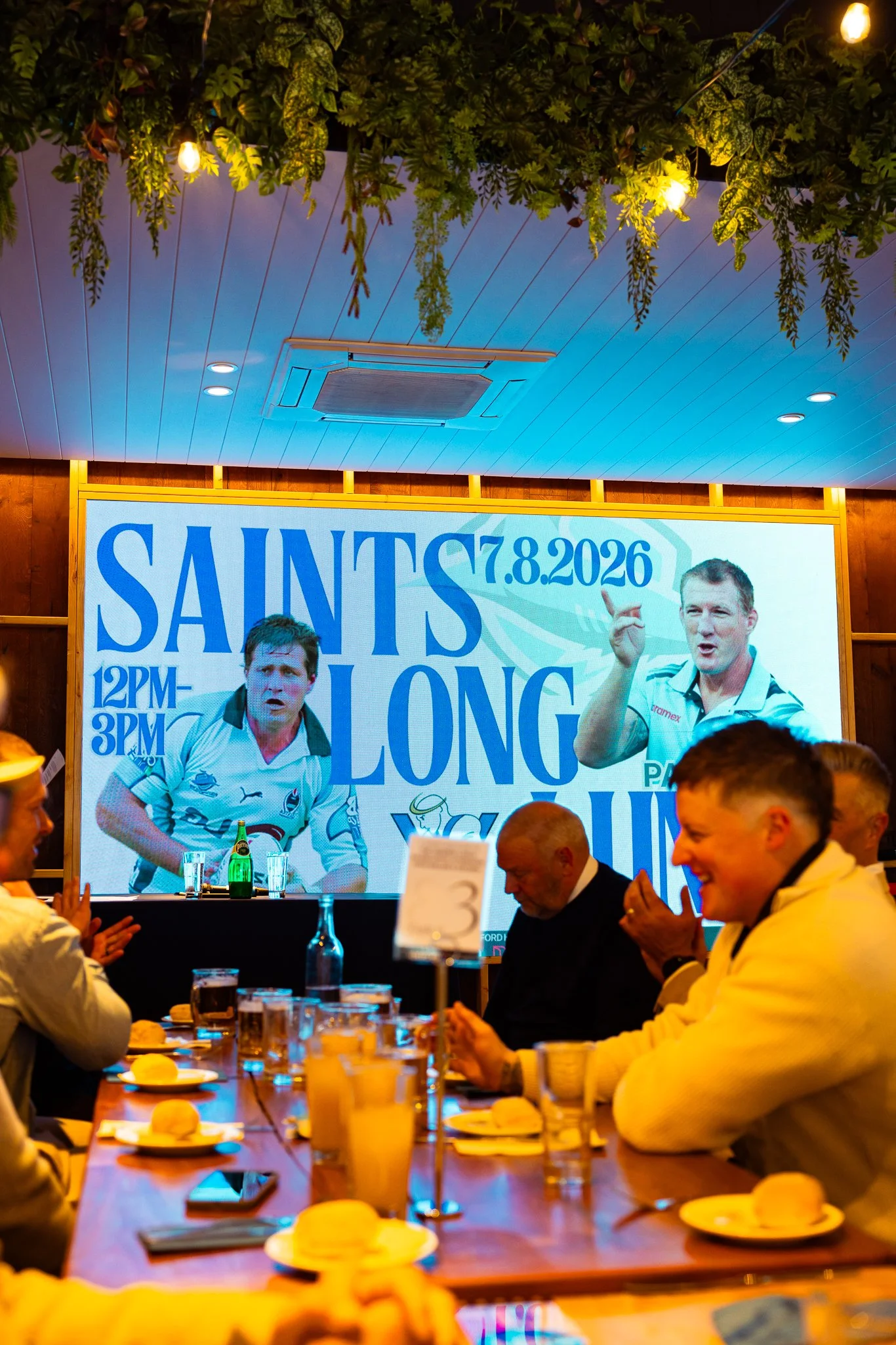 A restaurant or event space with people sitting at a long table eating and drinking. There is a large digital screen on the wall with a sports event advertisement, featuring rugby players and the date 7.8.2026.