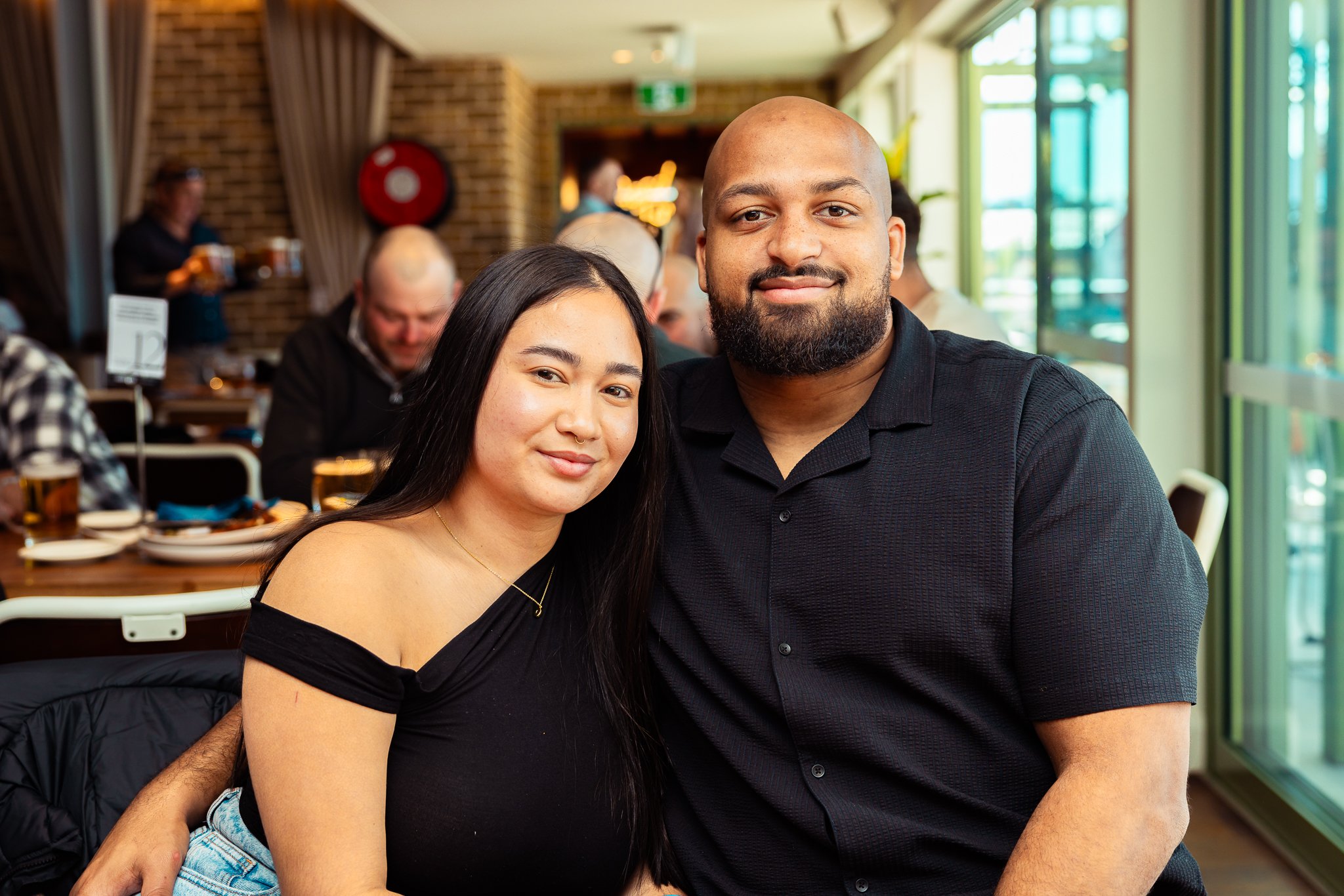 A young woman with long black hair and light skin, and a man with a beard and dark skin, pose together at a restaurant with large windows, brick walls, and other diners in the background.