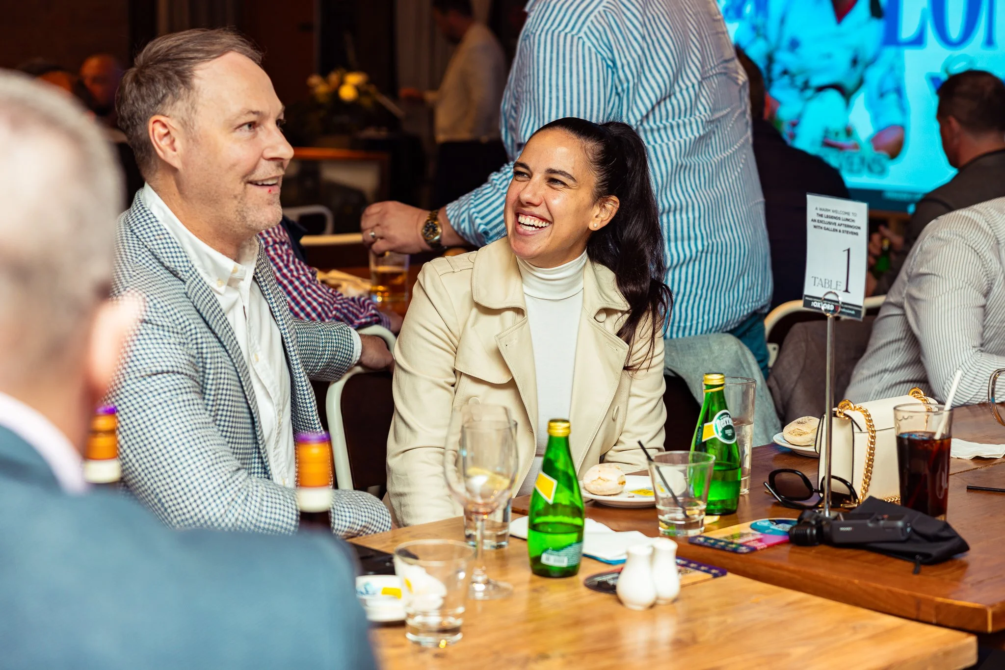 A group of people sitting at a table, smiling and talking in a lively social gathering, with drinks and snacks on the table.