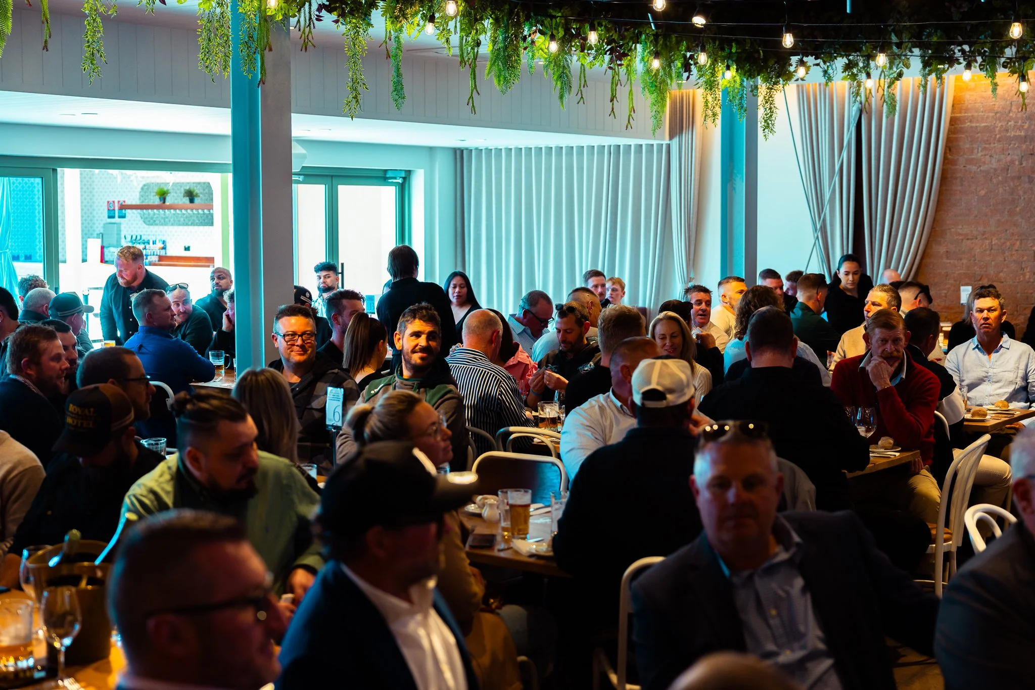 Photo of a large group of people sitting at tables in a restaurant or event space, engaged in conversation. The room is decorated with hanging greenery and string lights, with large windows and curtains in the background.