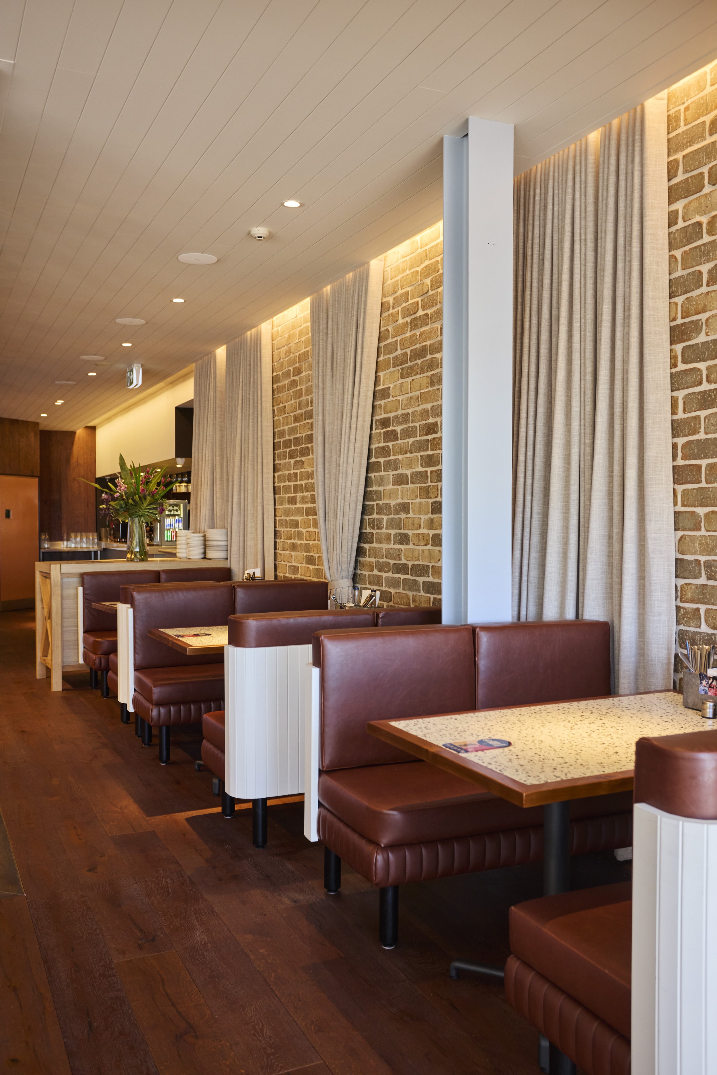Interior of a restaurant or cafe with brown leather booths, beige curtains, exposed brick walls, and a wooden floor. There is a table with a napkin holder, utensils, and a mobile phone on it, as well as a vase with flowers in the background.