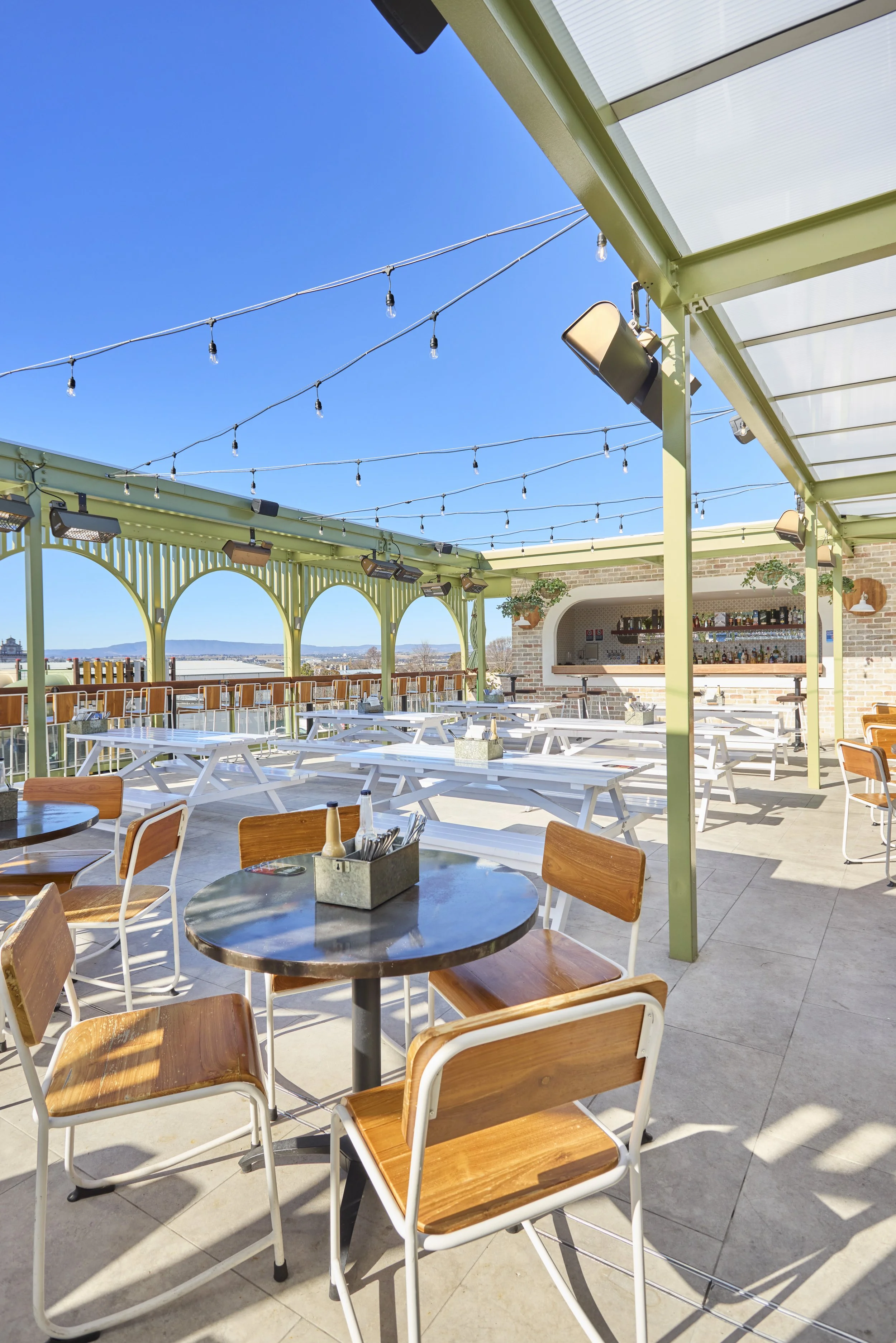 Empty outdoor rooftop bar area with tables, chairs, string lights, and a bar in the background against a blue sky.