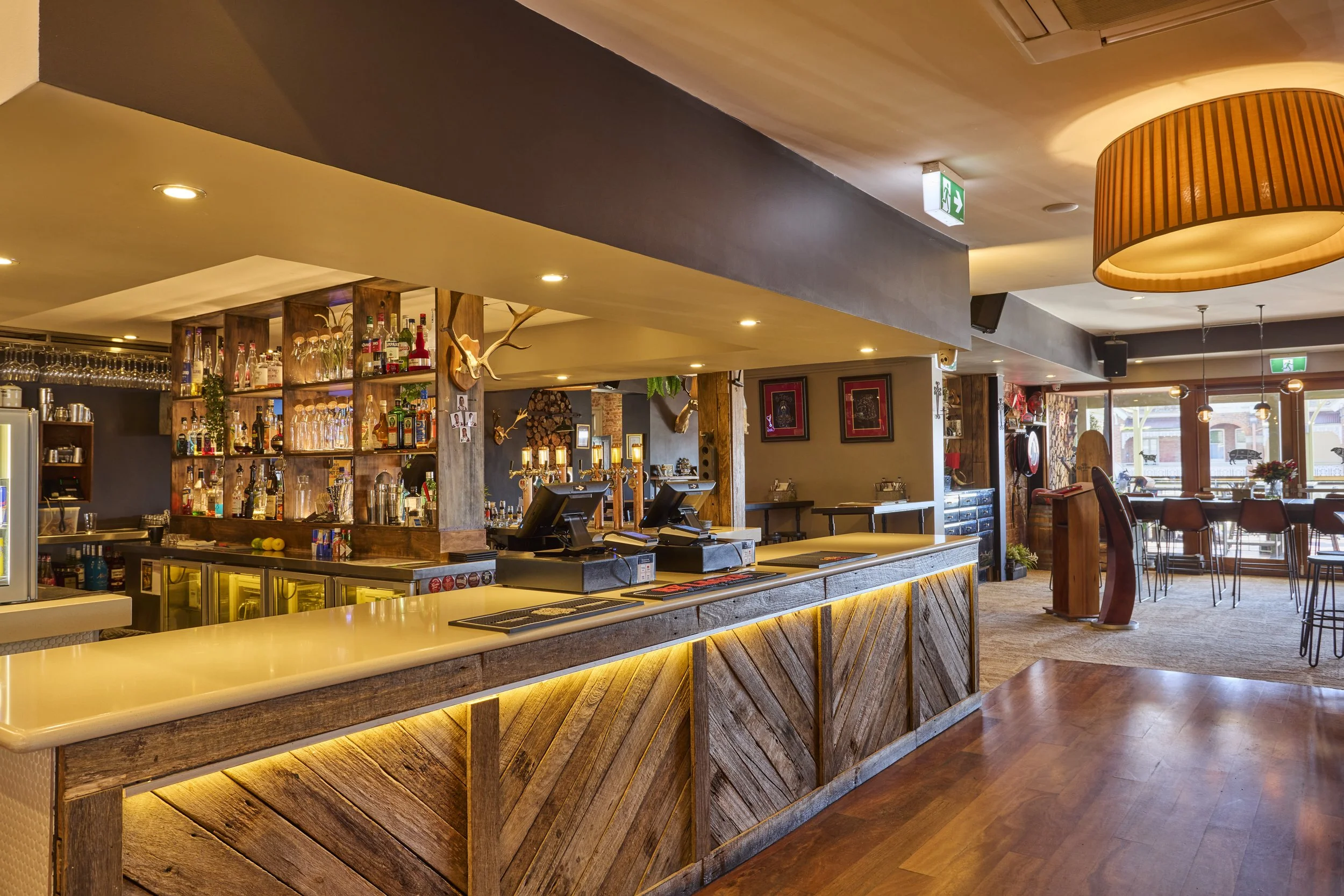 Interior of a bar or restaurant with a wooden counter, shelves with bottles and glasses, warm lighting, and modern decor.