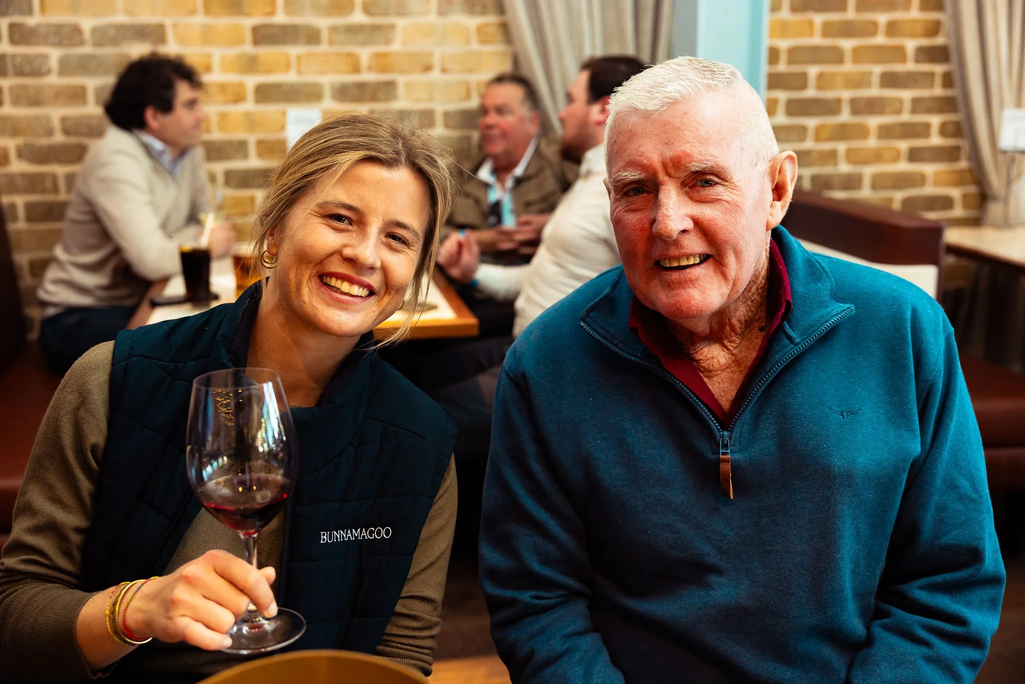 Two smiling people in the foreground, a woman holding a glass of red wine and an elderly man, with a group of people in the background at a social gathering in a brick-walled room.